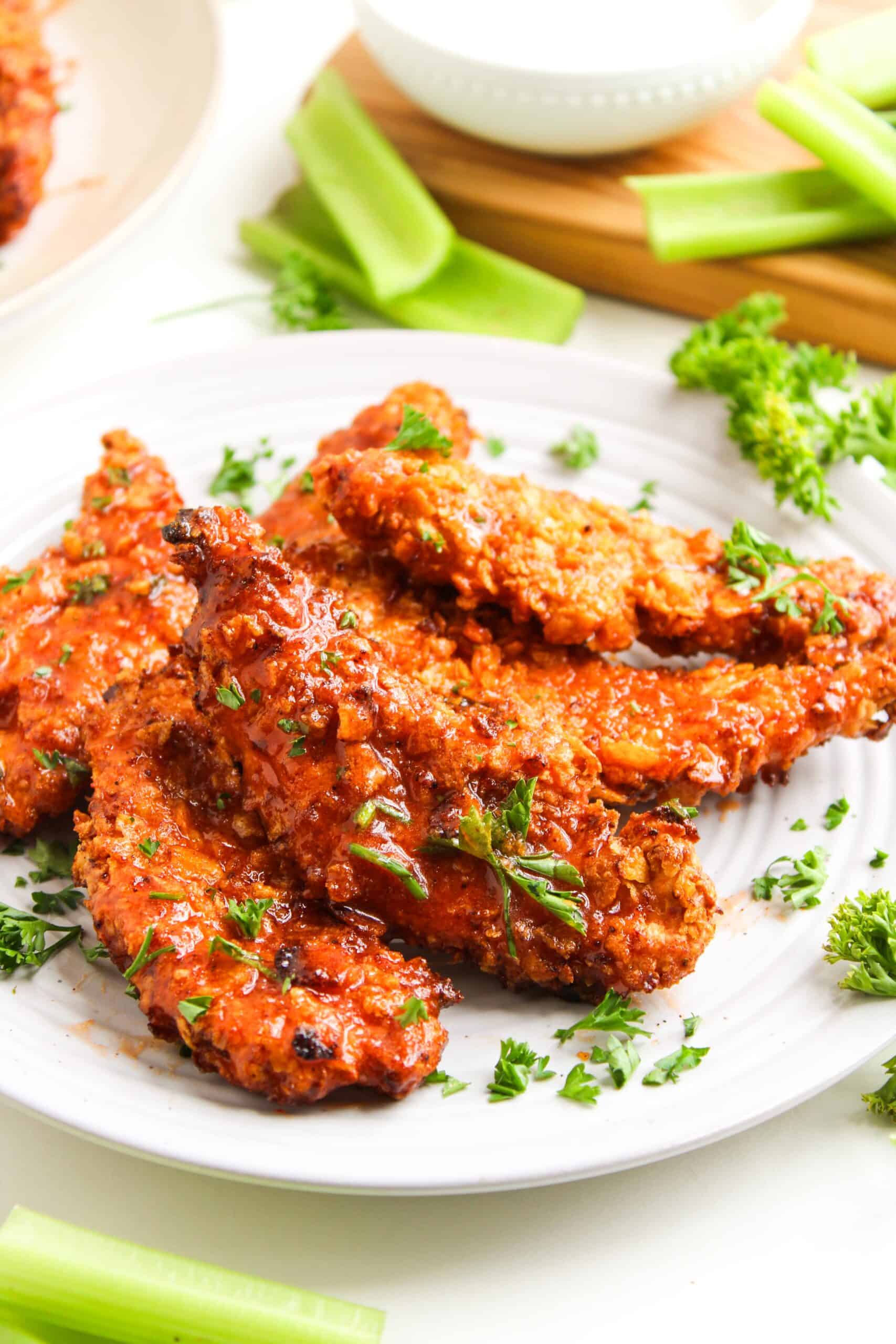 A plate of crispy Air Fryer Chicken tenders garnished with chopped parsley, served with celery sticks and a bowl of hot honey dipping sauce in the background.