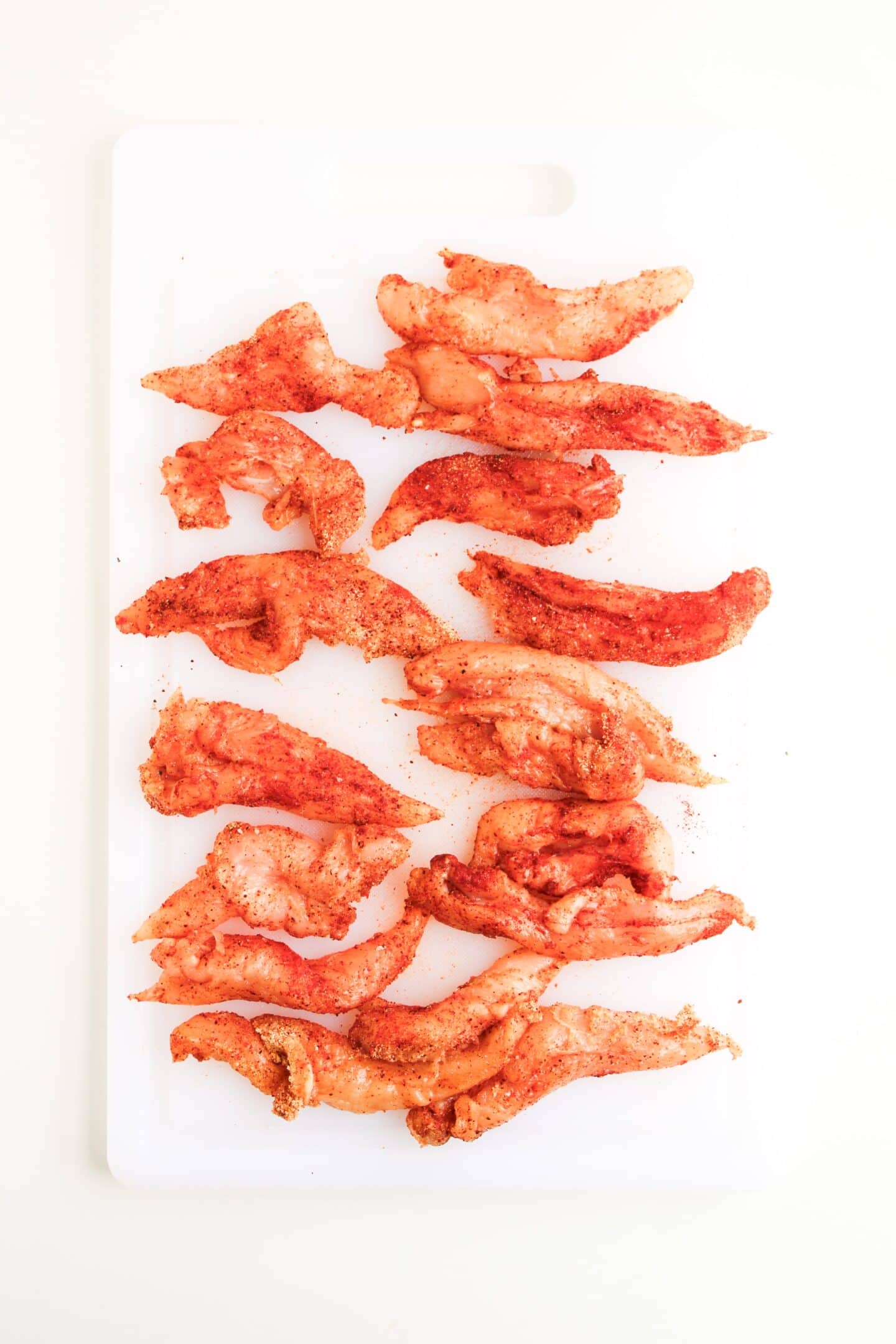 Raw, seasoned chicken strips arranged on a white cutting board, viewed from above, ready to become crispy Air Fryer Chicken Tenders on a plain white background.