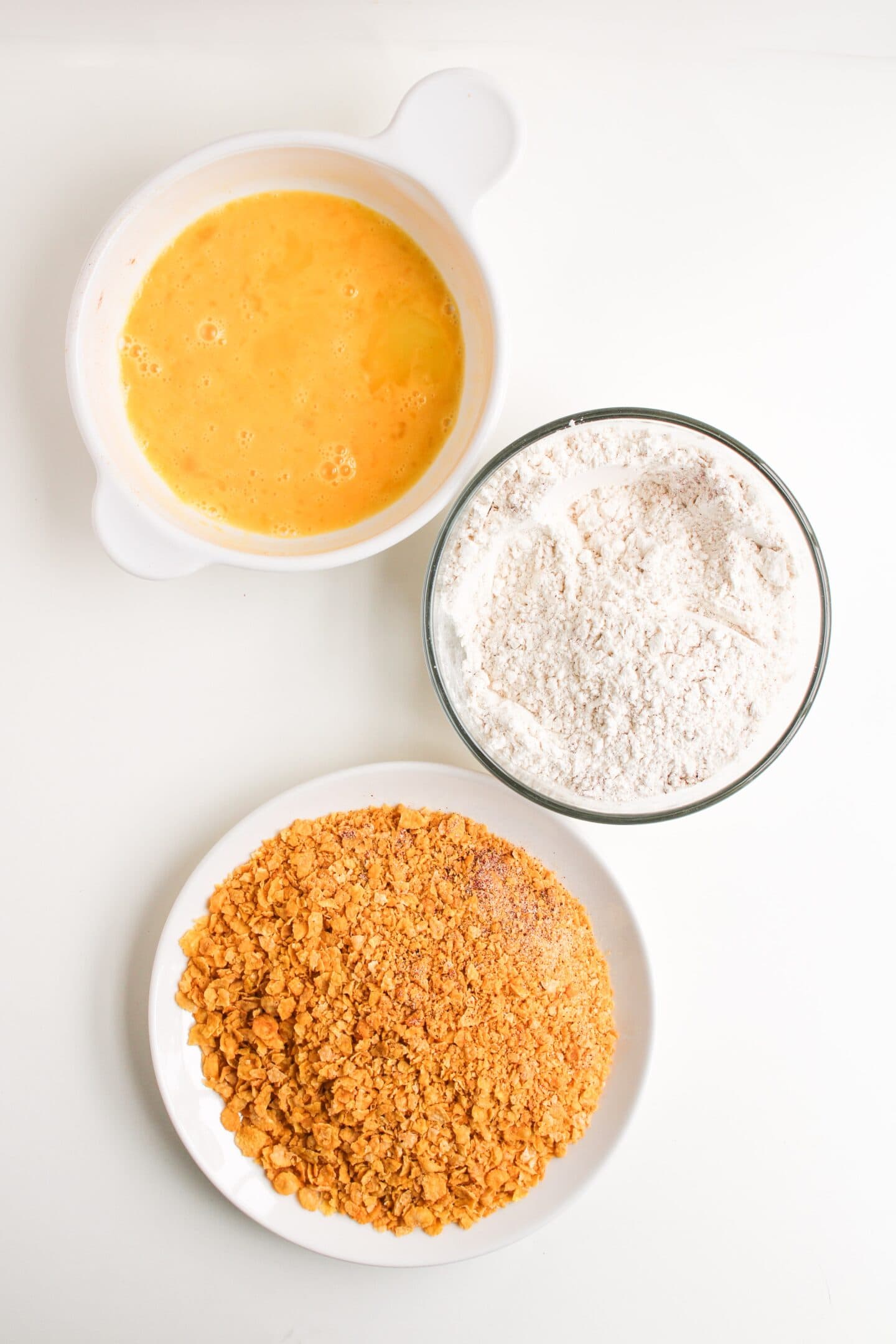 Three bowls on a white surface: one with beaten eggs, one with flour, and one with crushed cornflakes for making air fryer chicken tenders.