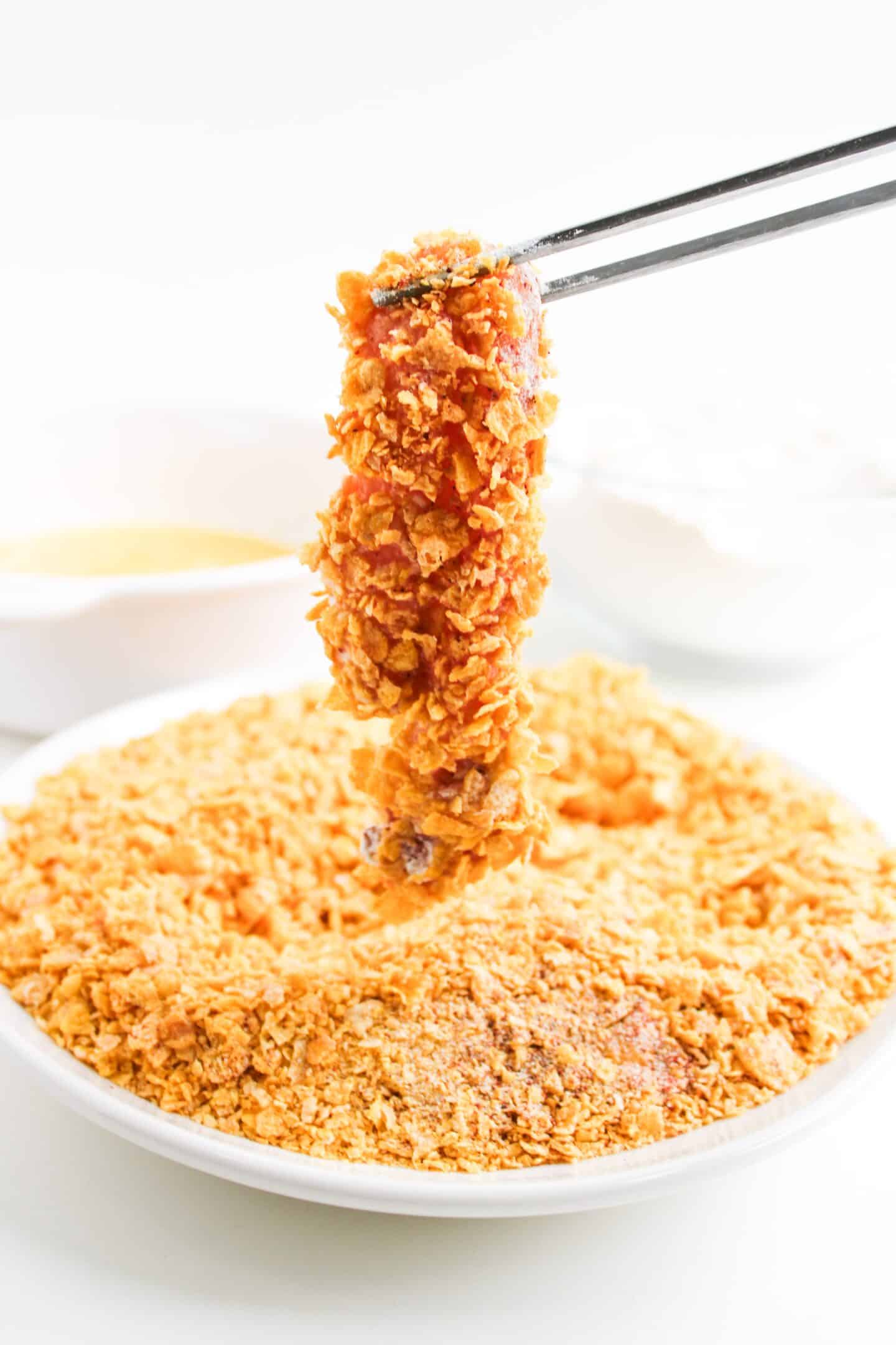 A piece of Air Fryer Chicken coated in crushed cornflakes is held with chopsticks above a plate filled with more crunchy cornflakes.