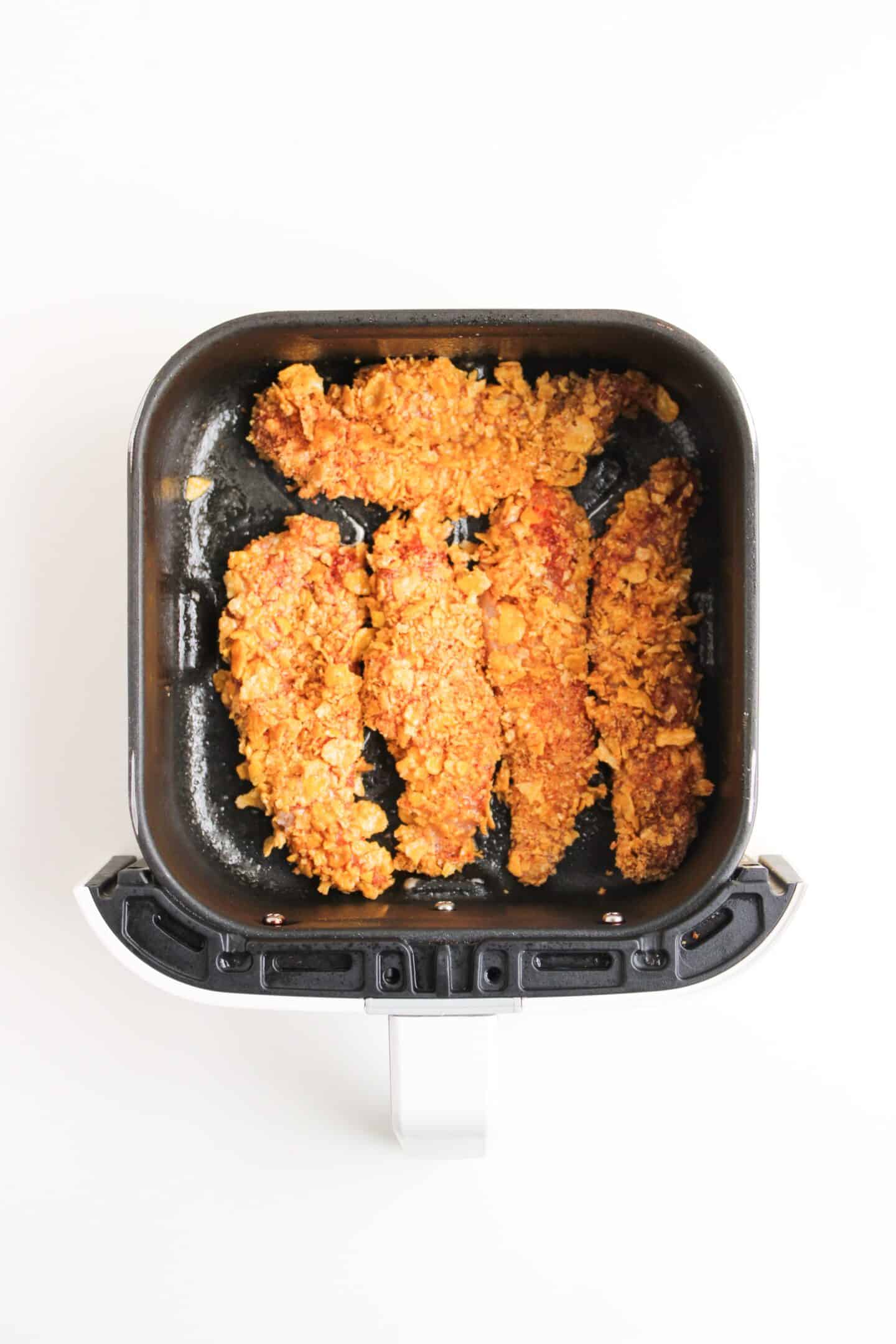 Four breaded chicken tenders are arranged inside an air fryer basket on a white background, ready to be enjoyed with a drizzle of hot honey.