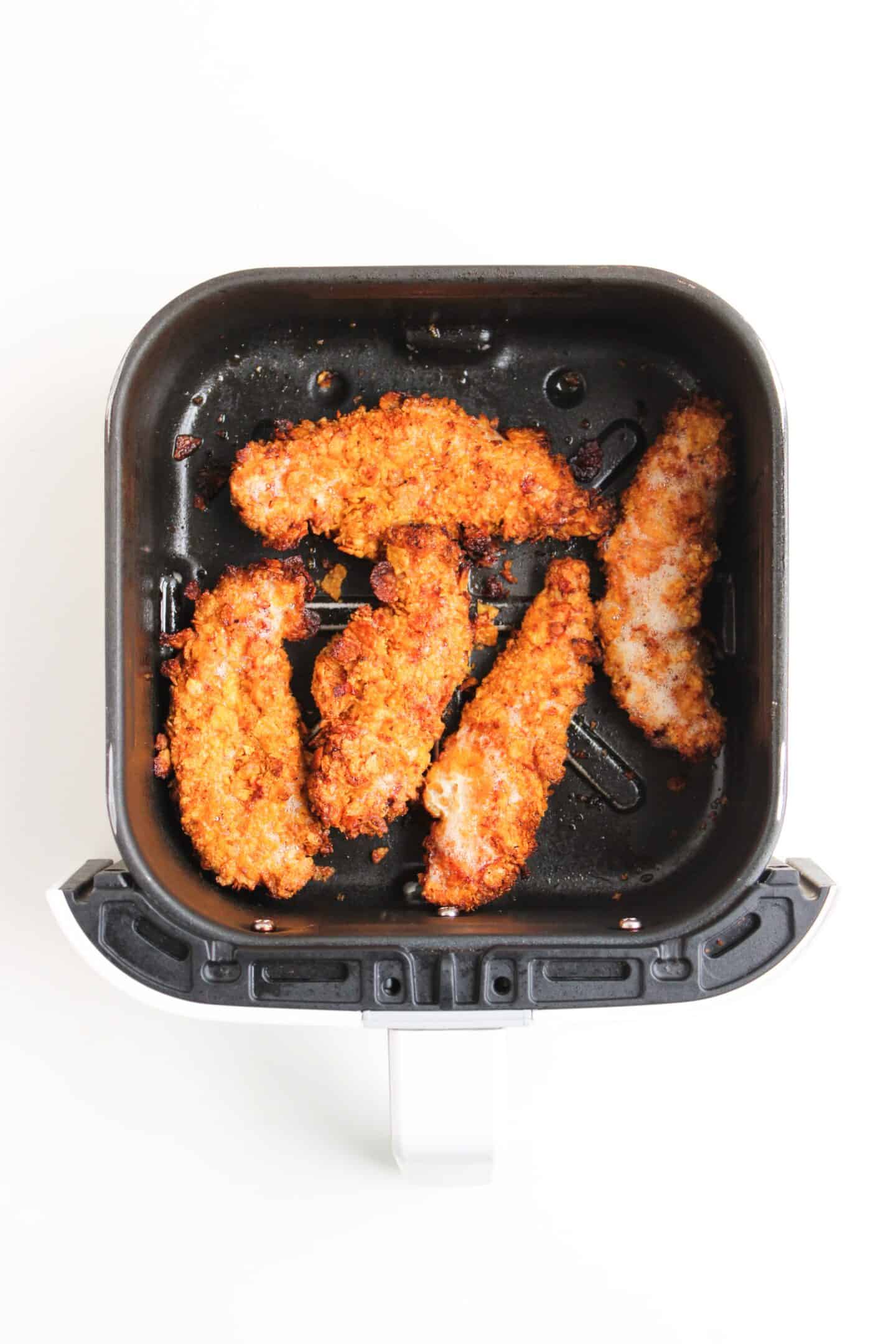 Five crispy Chicken Tenders are arranged in the basket of an Air Fryer, ready to be drizzled with hot honey, set against a white background.