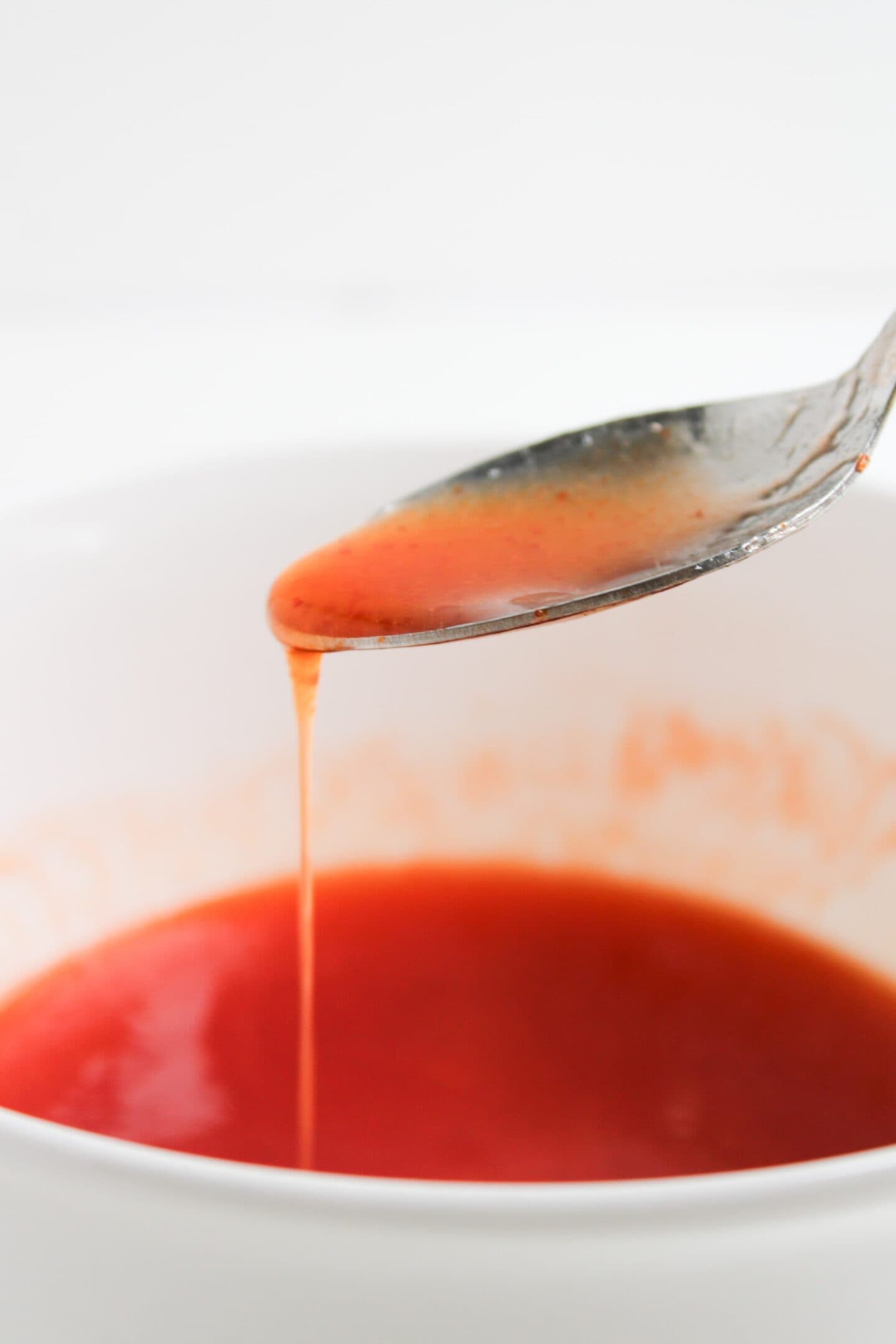 A metal spoon dripping red sauce—perfect for hot honey chicken tenders—into a white bowl filled with the same vibrant sauce.