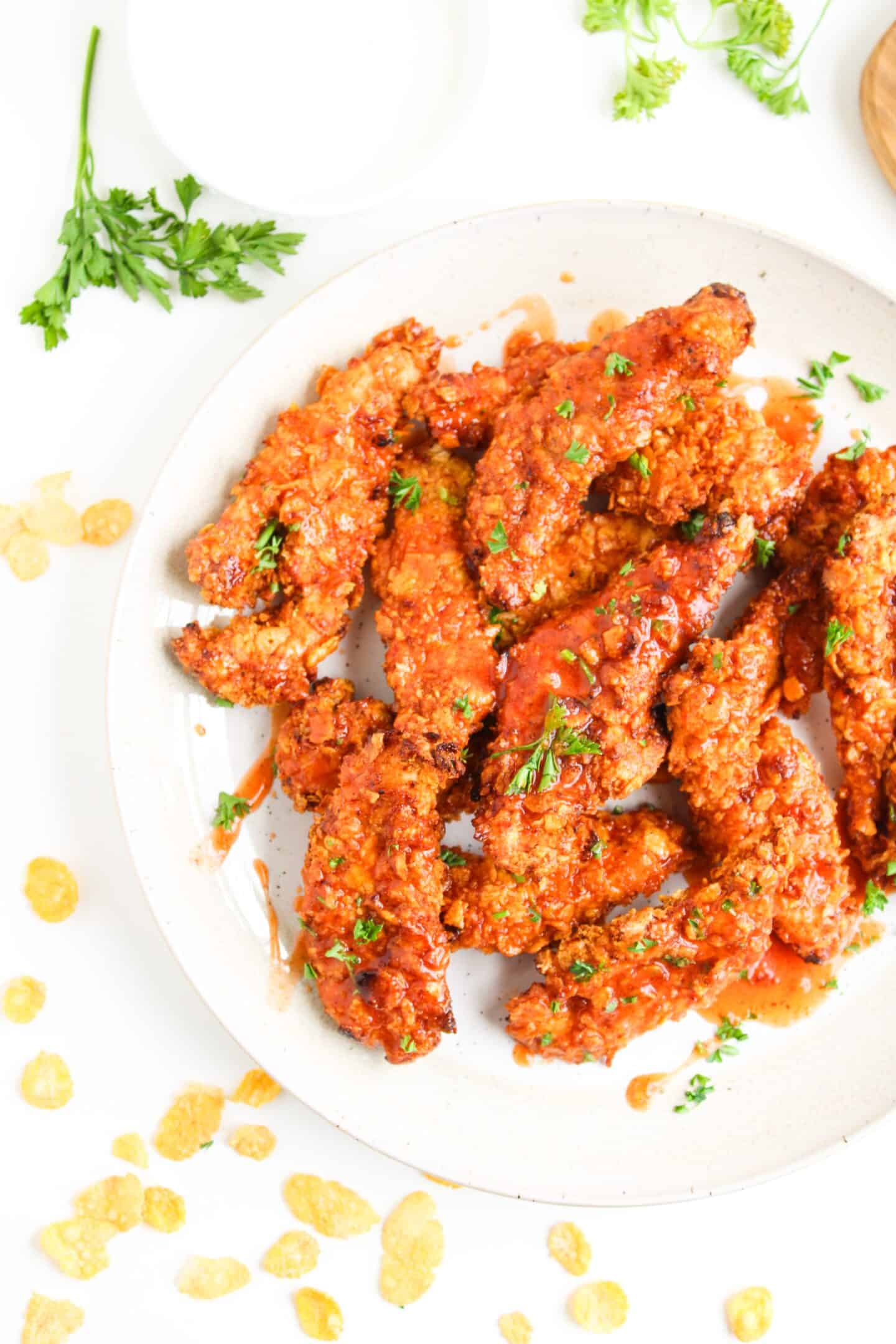 A plate of crispy, golden-brown Air Fryer Chicken Tenders coated in hot honey sauce and garnished with chopped parsley, with cornflakes and parsley scattered on the white surface nearby.