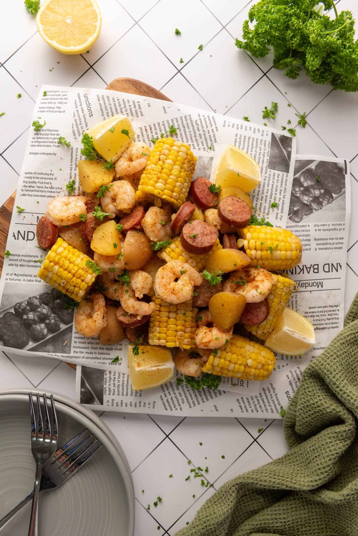 A classic Shrimp Boil platter—shrimp, sausage, corn on the cob, and potatoes—is served on newspaper with lemon wedges and parsley garnish. Plates, utensils, a lemon half, and parsley are nearby for this easy Instant Pot recipe.