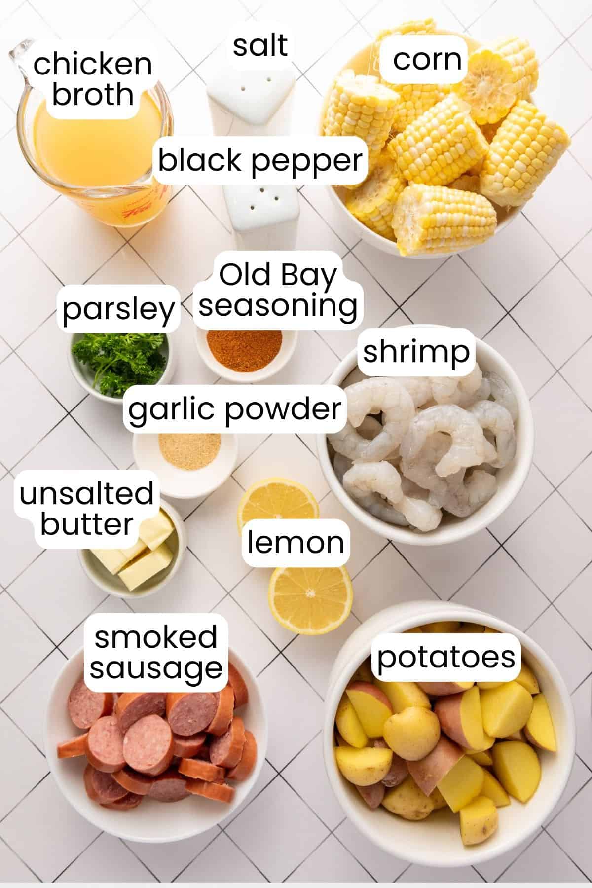Overhead view of ingredients for an Instant Pot shrimp boil, including corn, potatoes, shrimp, smoked sausage, lemon, butter, parsley, seasonings, and chicken broth on a white tiled surface.