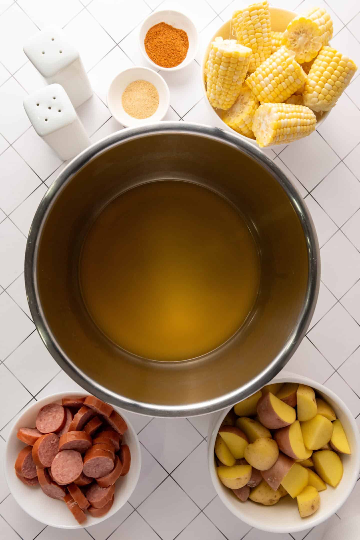 A large pot of broth is surrounded by bowls of sliced sausage, chopped potatoes, corn on the cob pieces, and small dishes of seasoning—everything you need for a classic Shrimp Boil recipe on a white grid-patterned surface.