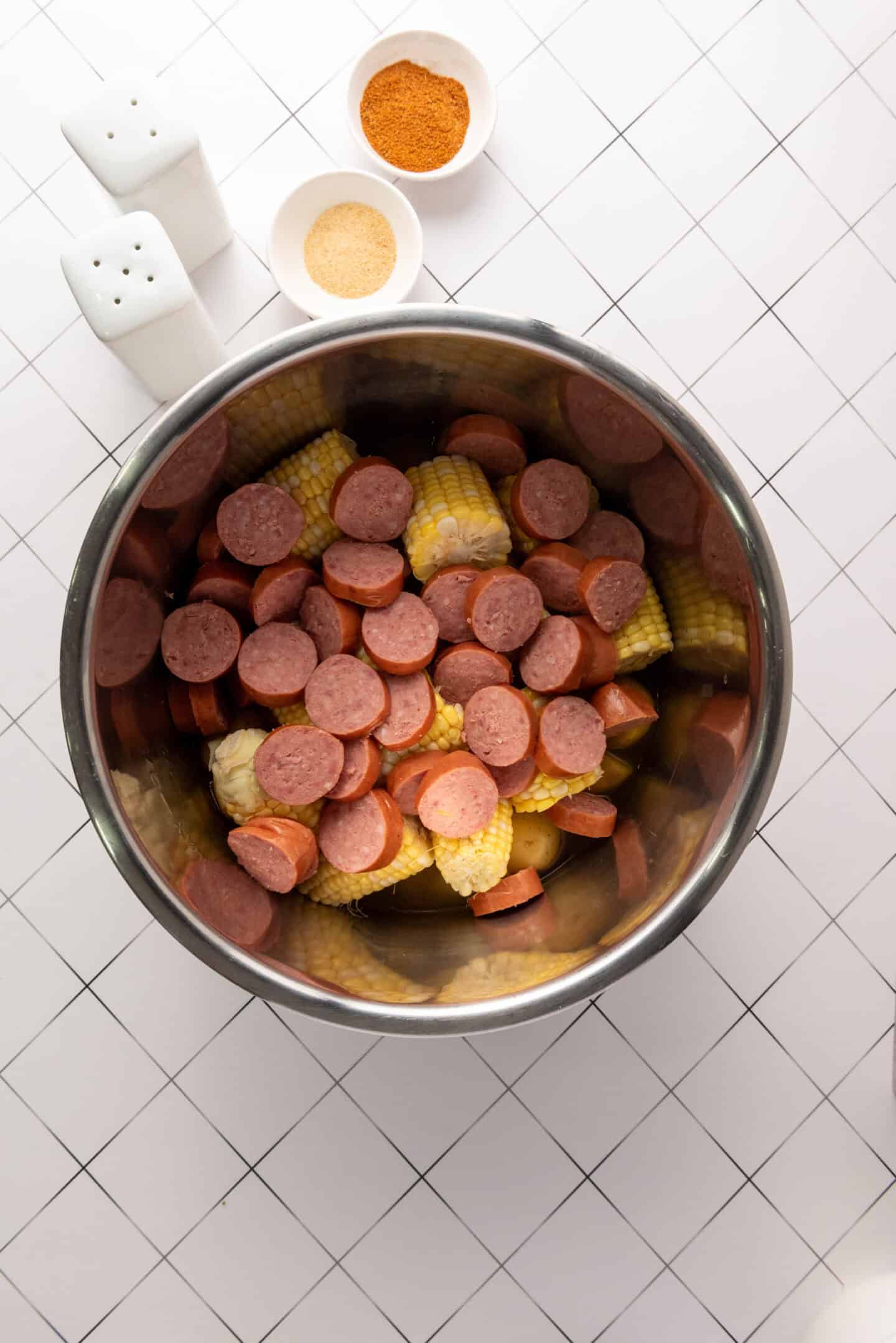 Sliced sausage, corn on the cob, and seasoning in a large metal pot, with two small bowls of spices and salt and pepper shakers on a tiled surface—perfect for an Instant Pot Shrimp Boil Recipe.