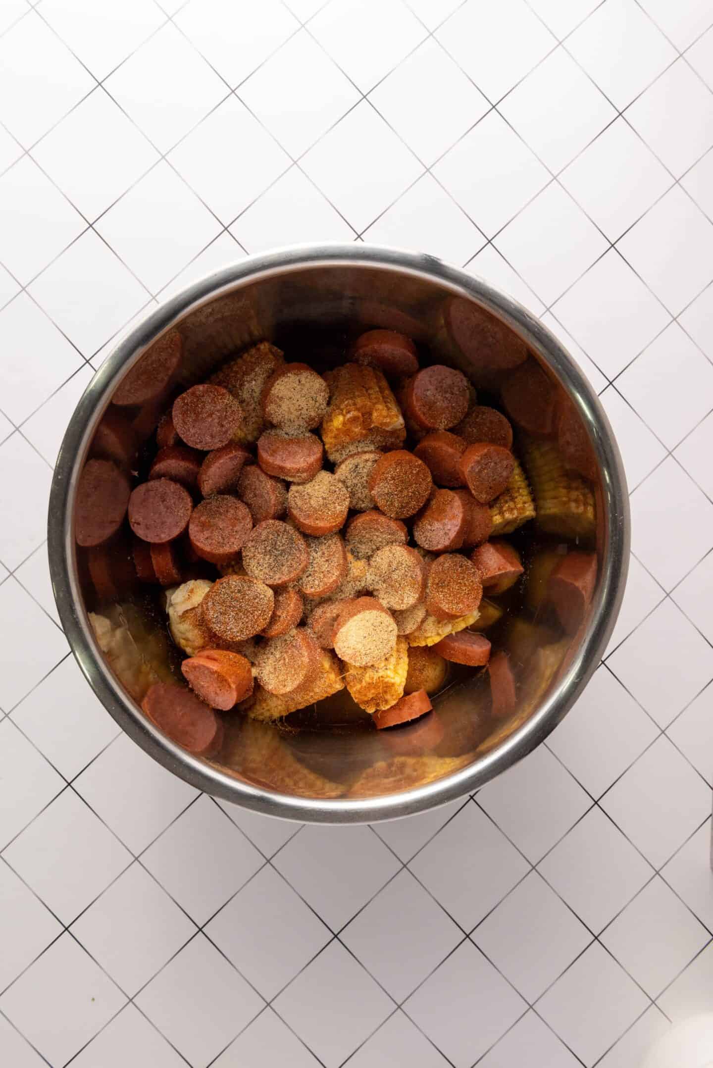 A metal bowl filled with sliced sausage, corn on the cob pieces, and seasonings, this easy Instant Pot Shrimp Boil recipe sits ready on a white tiled surface.