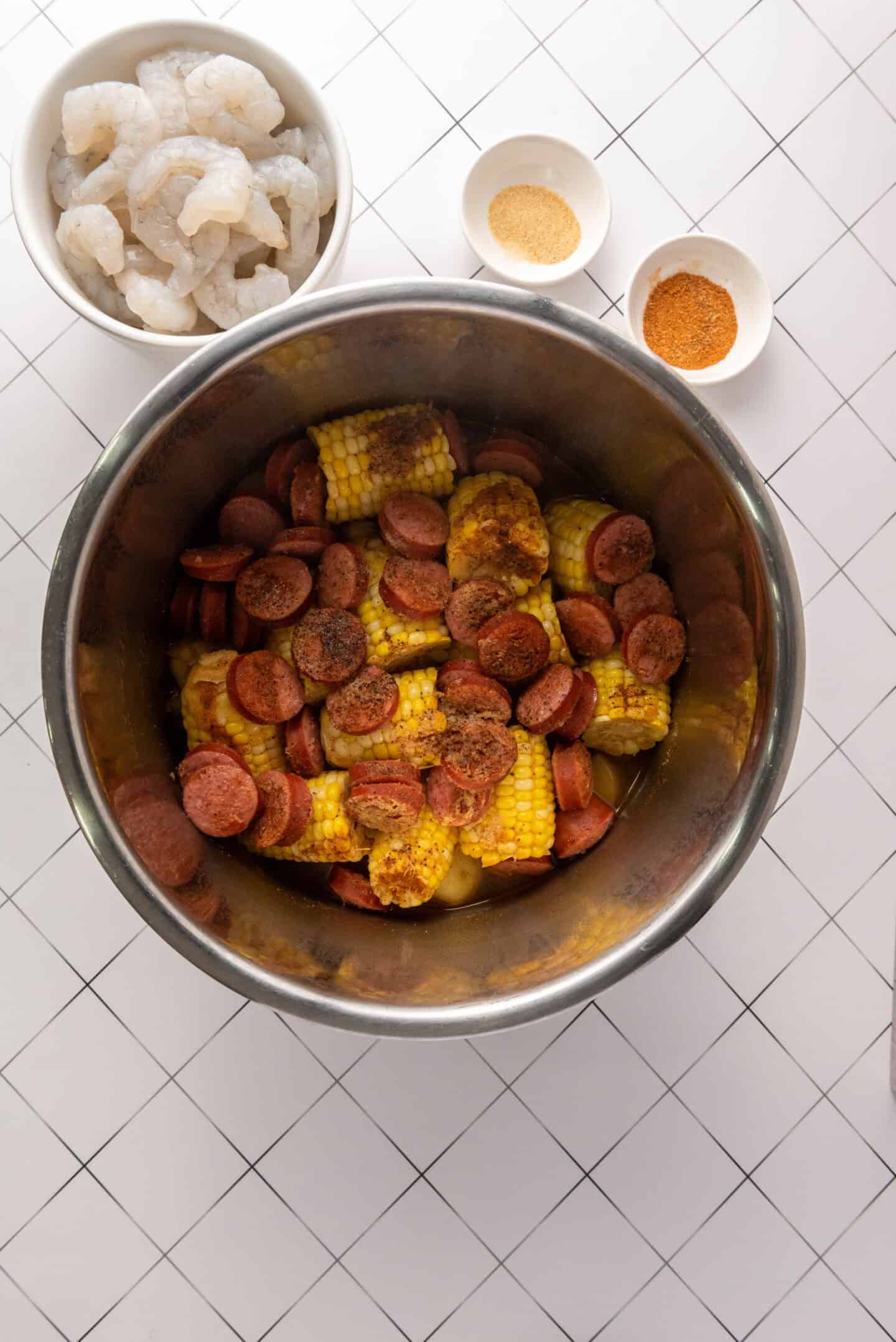 A metal bowl with sliced sausage, corn on the cob, and seasoning sits ready for a Shrimp Boil recipe. Nearby are bowls of raw shrimp and two types of powdered seasoning, all arranged on a white tiled surface.
