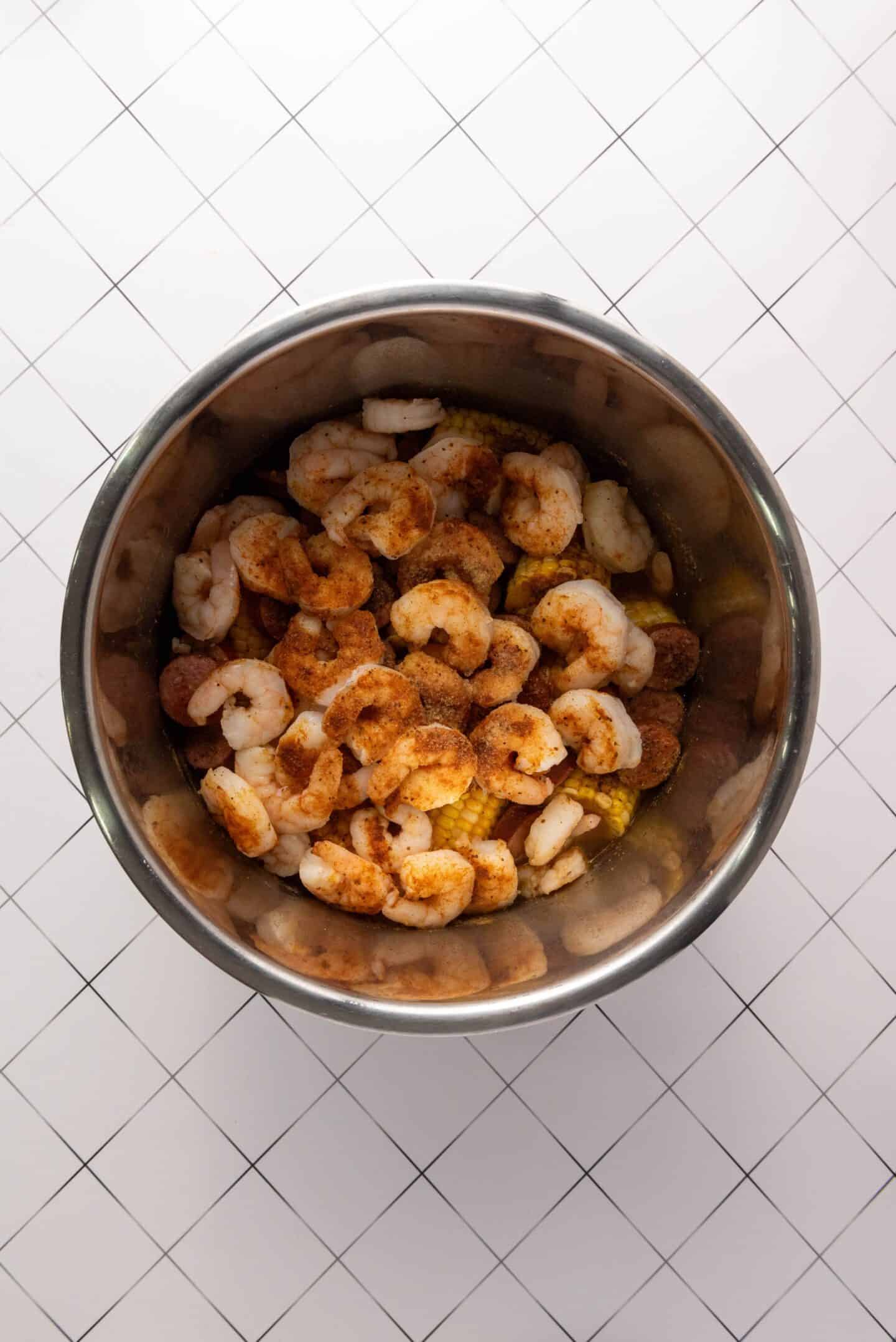 A metal bowl filled with raw shrimp seasoned with spices, perfect for a Shrimp Boil recipe, placed on a white tiled surface with a grid pattern.