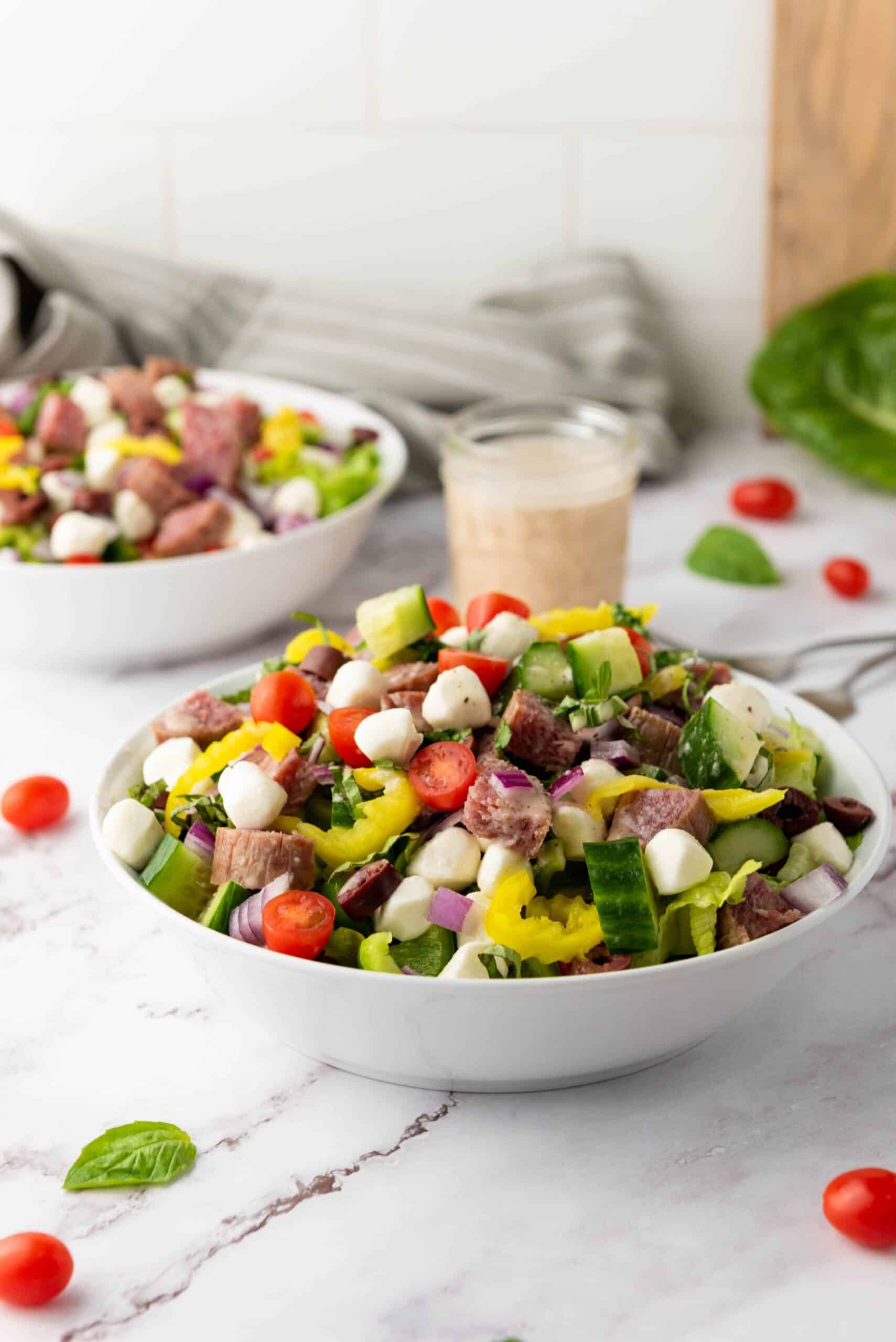 A bowl of Italian Chopped Salad with chunks of steak, cherry tomatoes, cucumbers, mozzarella balls, pepperoncini, and red onions sits on a marble surface, accompanied by zesty dressing in the background.