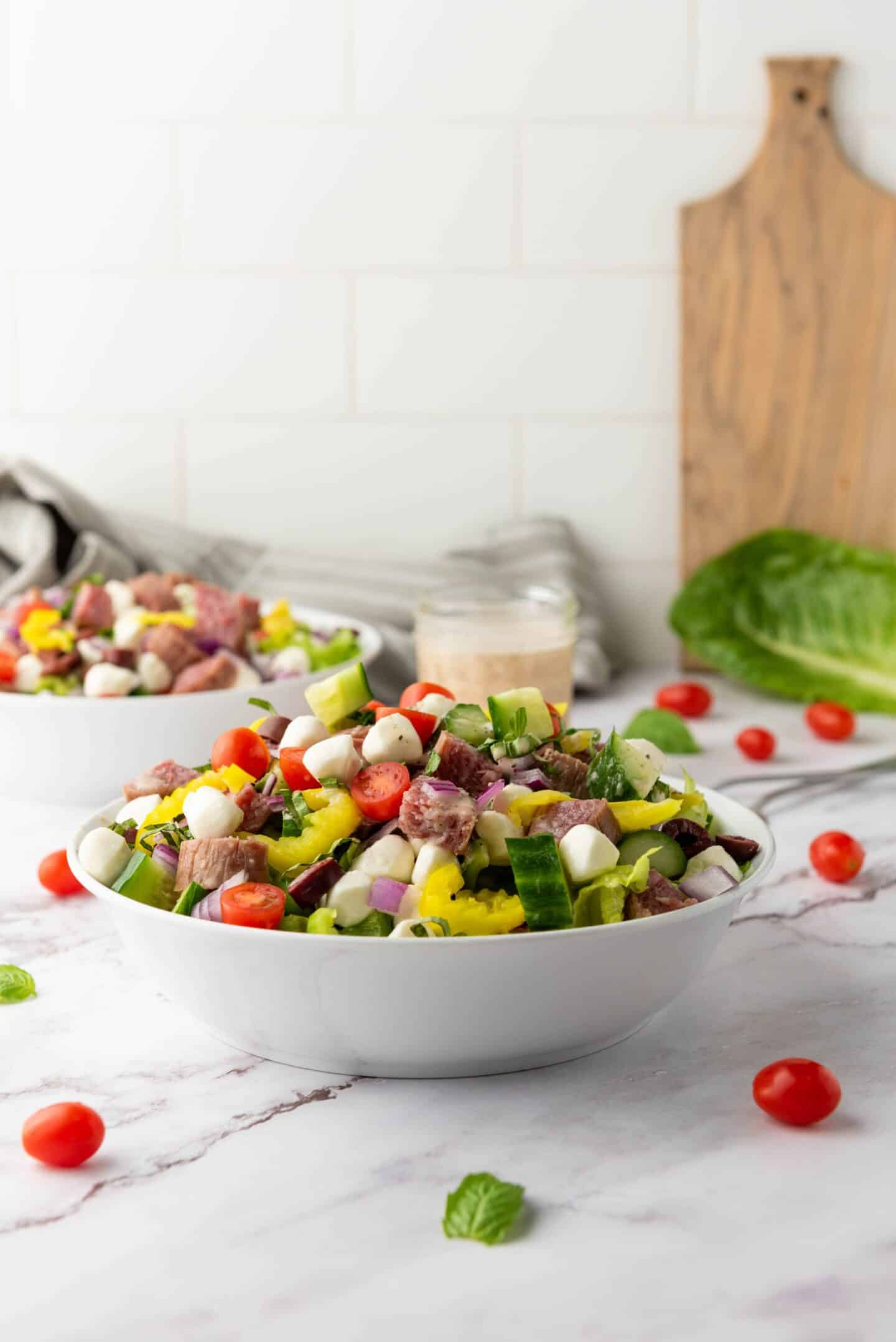A bowl of Italian Chopped Salad with cucumbers, tomatoes, mozzarella balls, red onion, peppers, and meat pieces sits on a marble countertop, with fresh salad ingredients and a zesty dressing in the background.