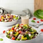 A bowl of Italian Chopped Salad featuring lettuce, cherry tomatoes, cucumber, mozzarella balls, red onion, and steak chunks, with a glass of zesty dressing in the background.