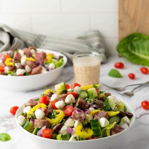 A bowl of Italian Chopped Salad featuring lettuce, cherry tomatoes, cucumber, mozzarella balls, red onion, and steak chunks, with a glass of zesty dressing in the background.