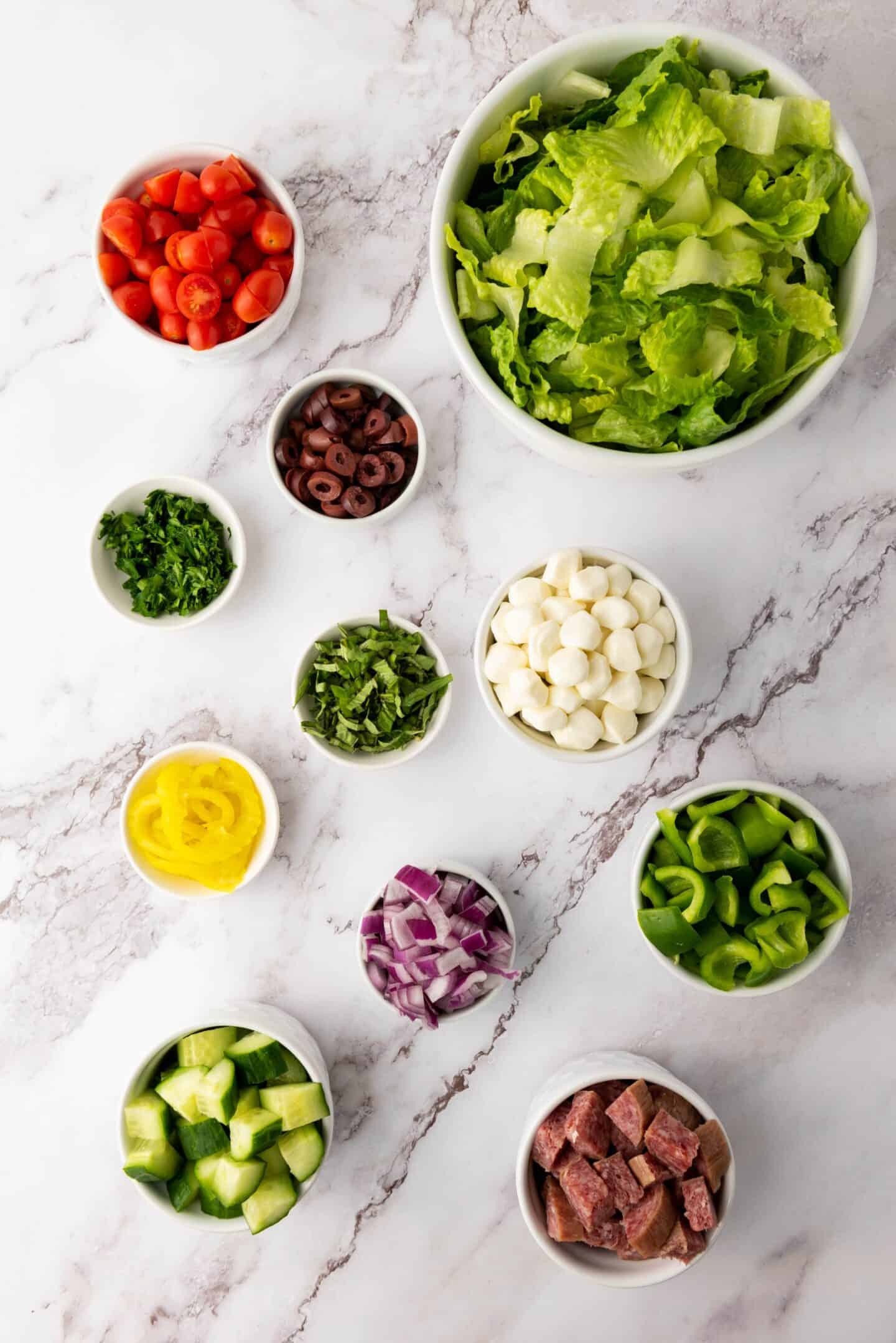 Assorted salad ingredients in bowls, including lettuce, cherry tomatoes, olives, mozzarella balls, cucumbers, peppers, red onion, herbs, and meat, arranged on a marble surface for a classic Italian Chopped Salad with zesty dressing.