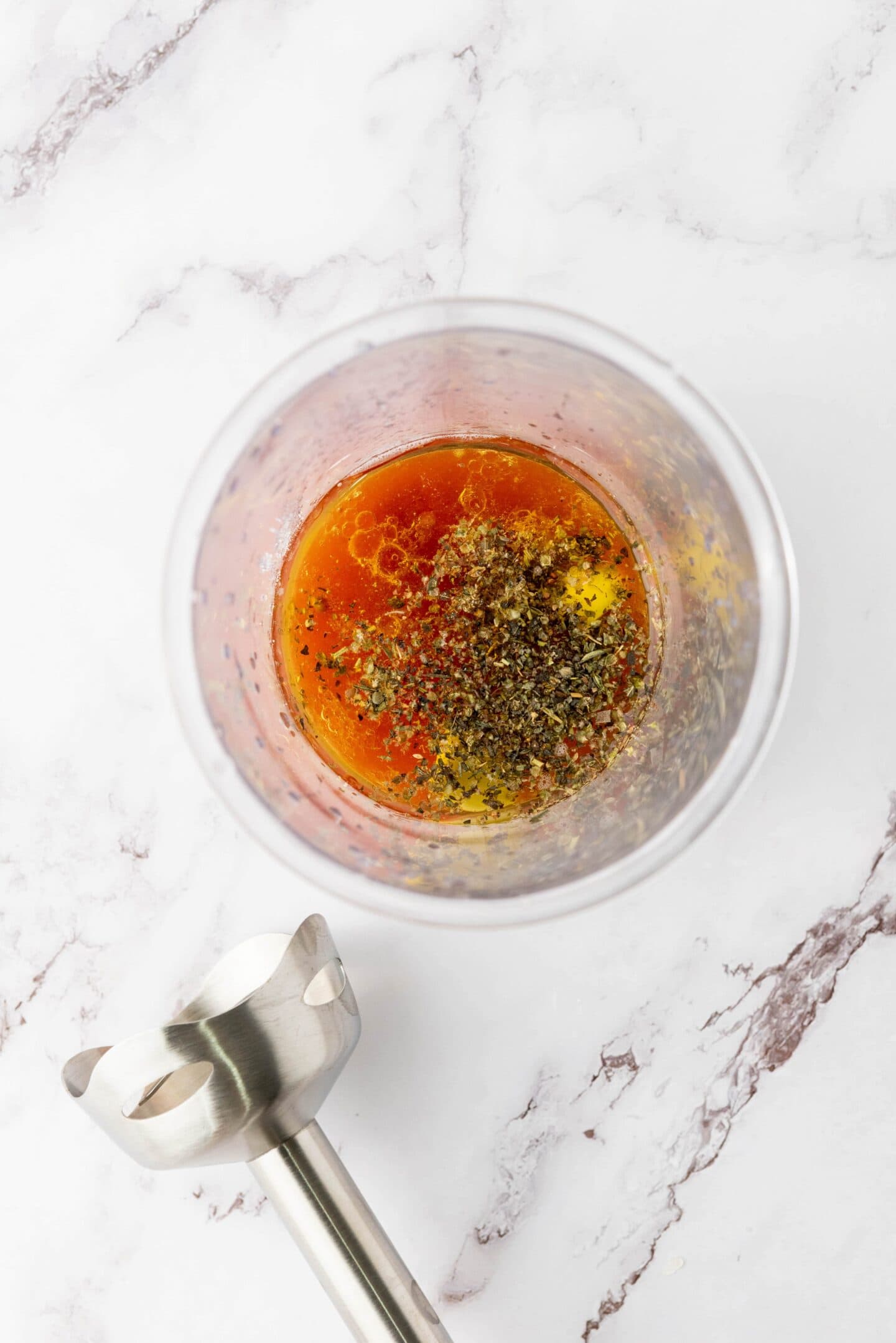 A glass container with oil, vinegar, herbs, and spices sits on a marble surface next to an immersion blender—perfect for whipping up a zesty dressing for your favorite Italian chopped salad.