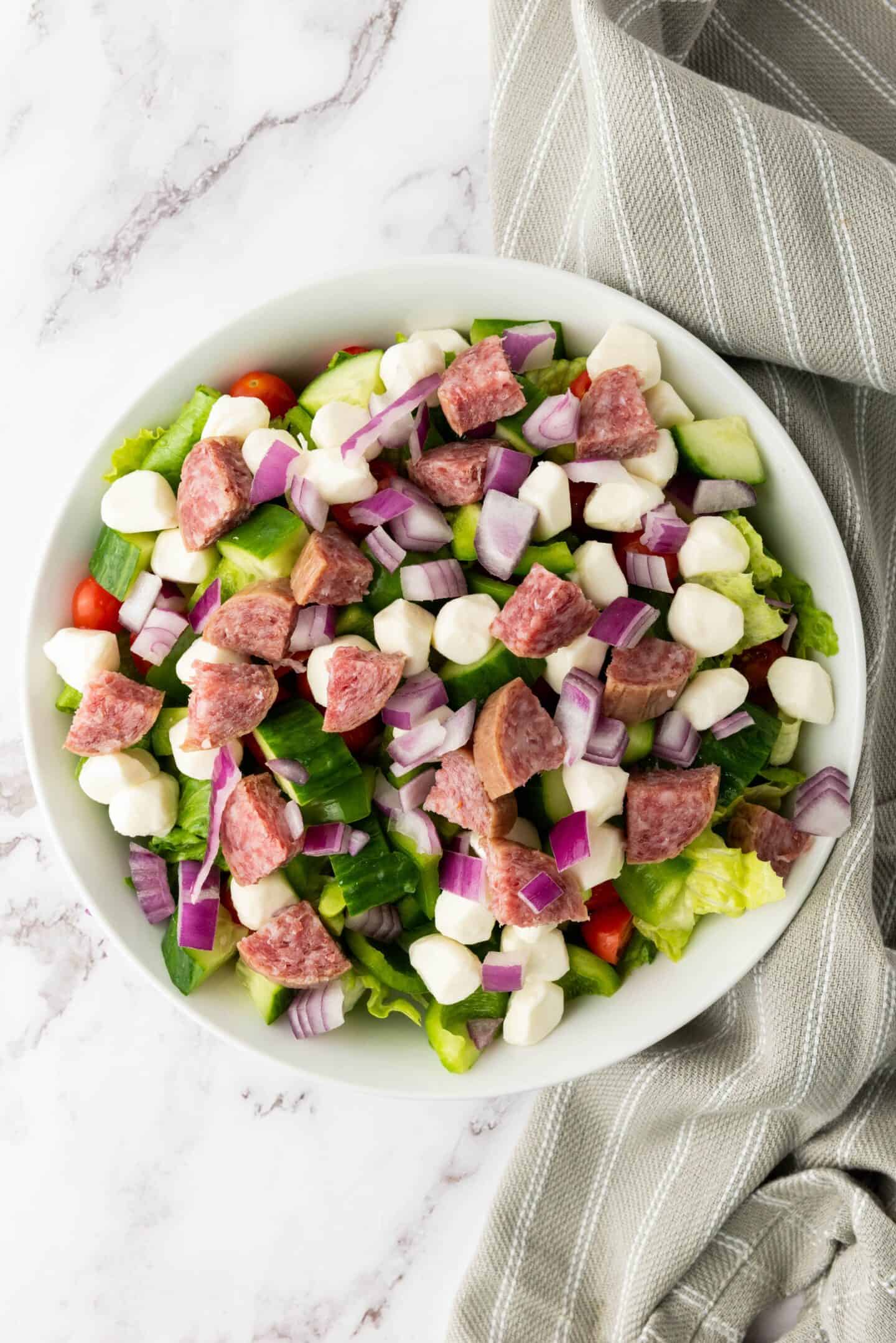 A bowl of Italian Chopped Salad with chopped lettuce, cherry tomatoes, salami slices, mozzarella balls, diced red onions, and cucumber sits on a marble surface beside a gray-striped cloth.