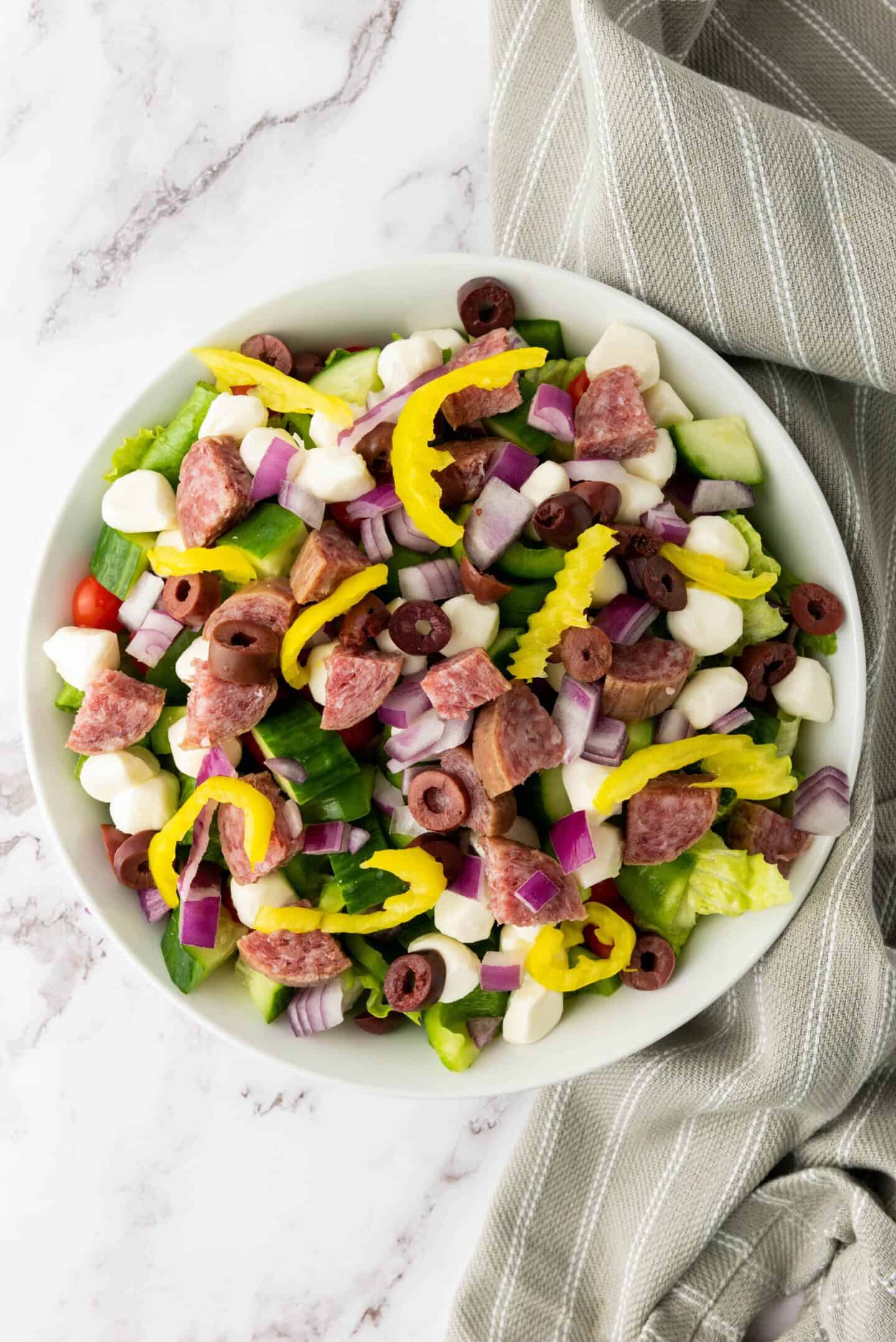 A bowl of Italian Chopped Salad with lettuce, salami, mozzarella balls, olives, red onions, banana peppers, cucumber, and grape tomatoes on a marble surface with a striped gray napkin.