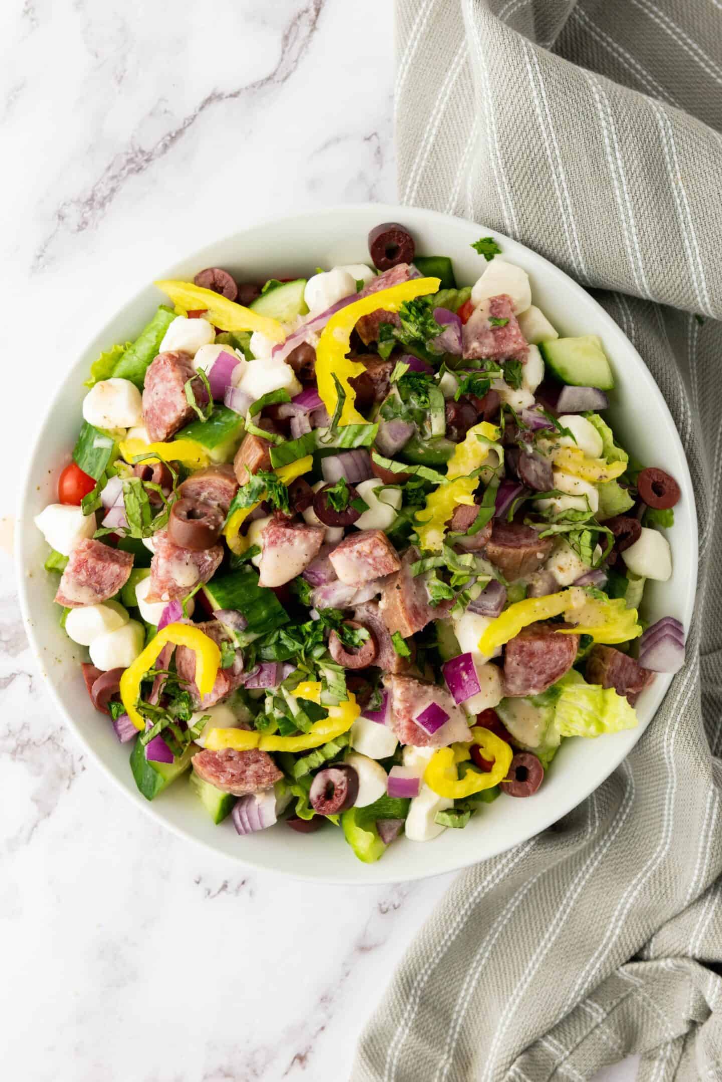 A bowl of Italian Chopped Salad with sliced sausage, lettuce, mozzarella balls, olives, onions, pepper rings, and chopped herbs sits on a marble surface with a striped napkin and zesty dressing.