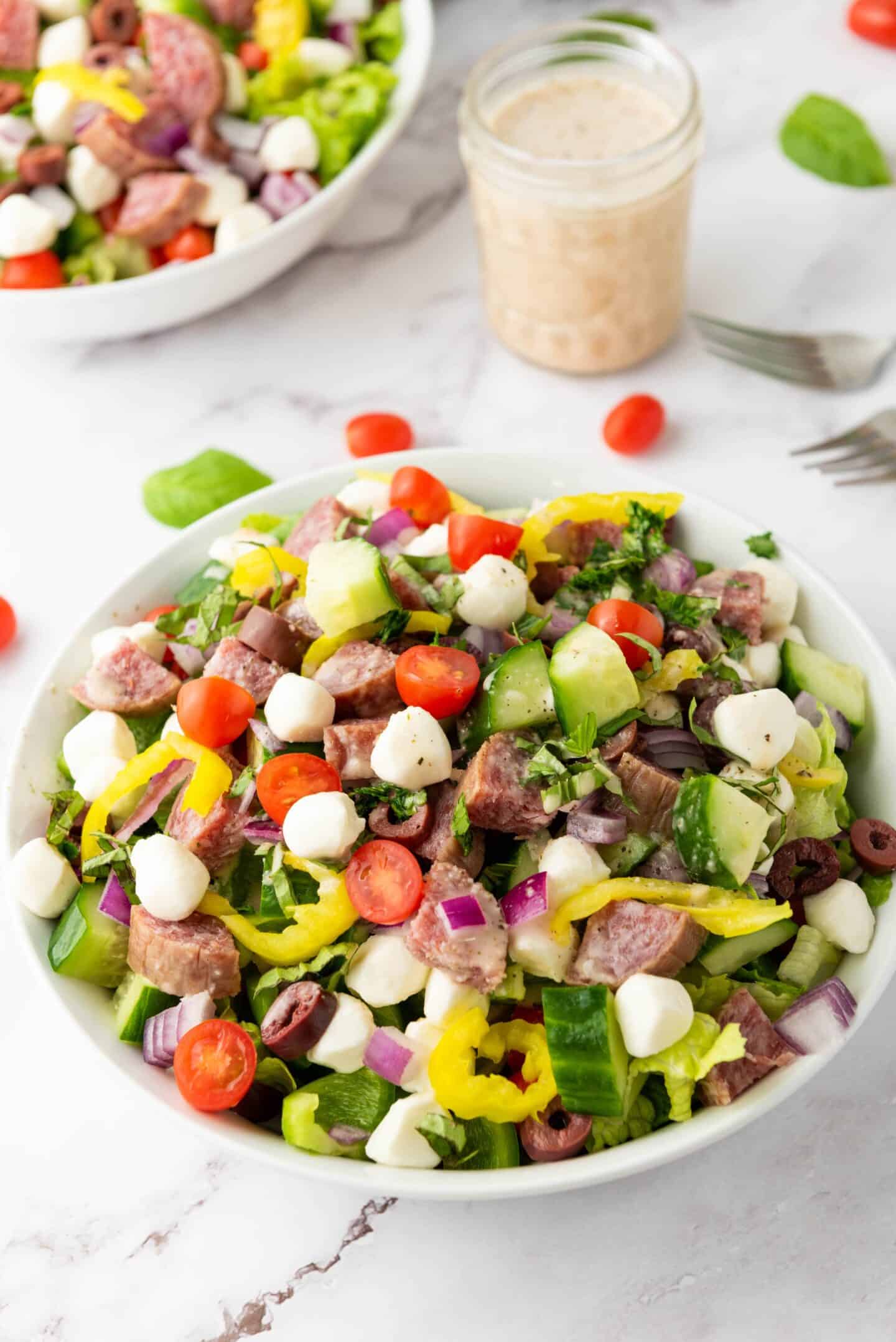 A bowl of Italian Chopped Salad with cherry tomatoes, mozzarella balls, salami, cucumbers, red onions, peppers, and olives. A jar of zesty dressing and another salad bowl are in the background.