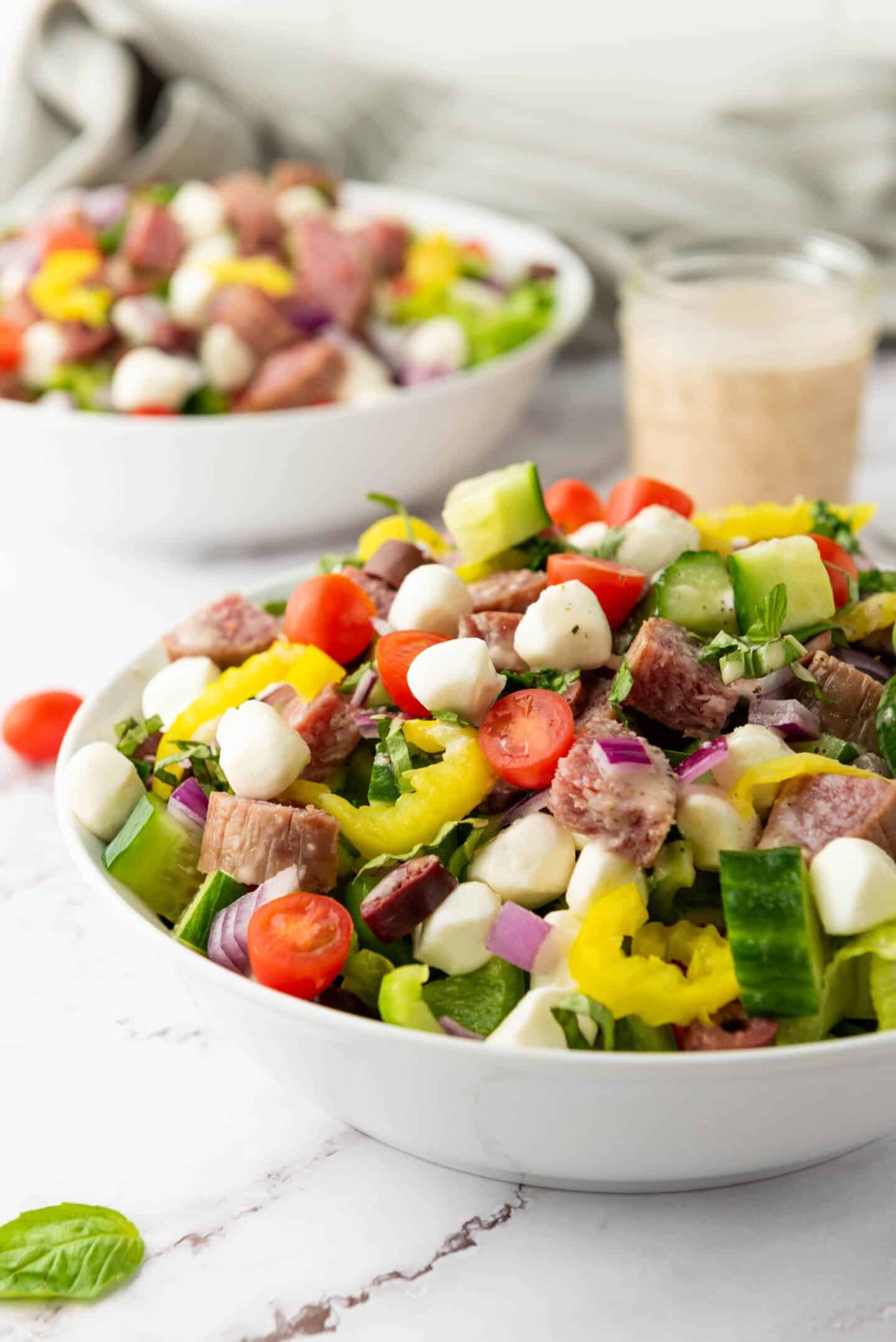 A bowl of Italian Chopped Salad with sliced salami, cherry tomatoes, mozzarella balls, cucumbers, red onions, and pepperoncini peppers, with a jar of zesty dressing in the background.