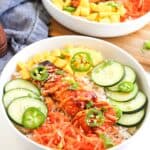 Two white bowls filled with Spicy Salmon Bowls ingredients: rice, grilled salmon, shredded carrots, sliced cucumber, jalapeño, mango, and green onions, all garnished with sesame seeds.