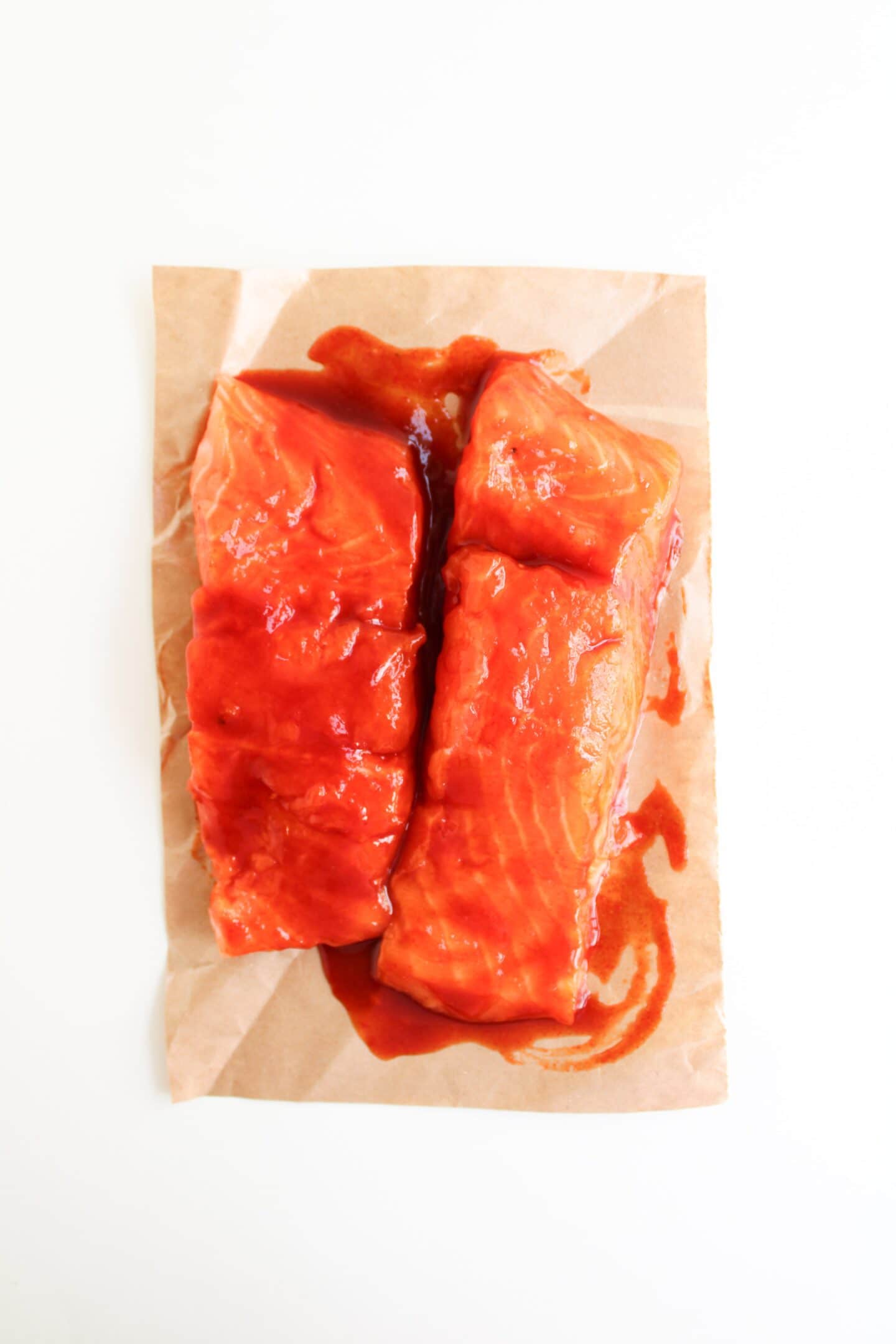 Two raw salmon fillets coated in a red marinade are placed on brown parchment paper against a white background, perfect for making Spicy Salmon Bowls.