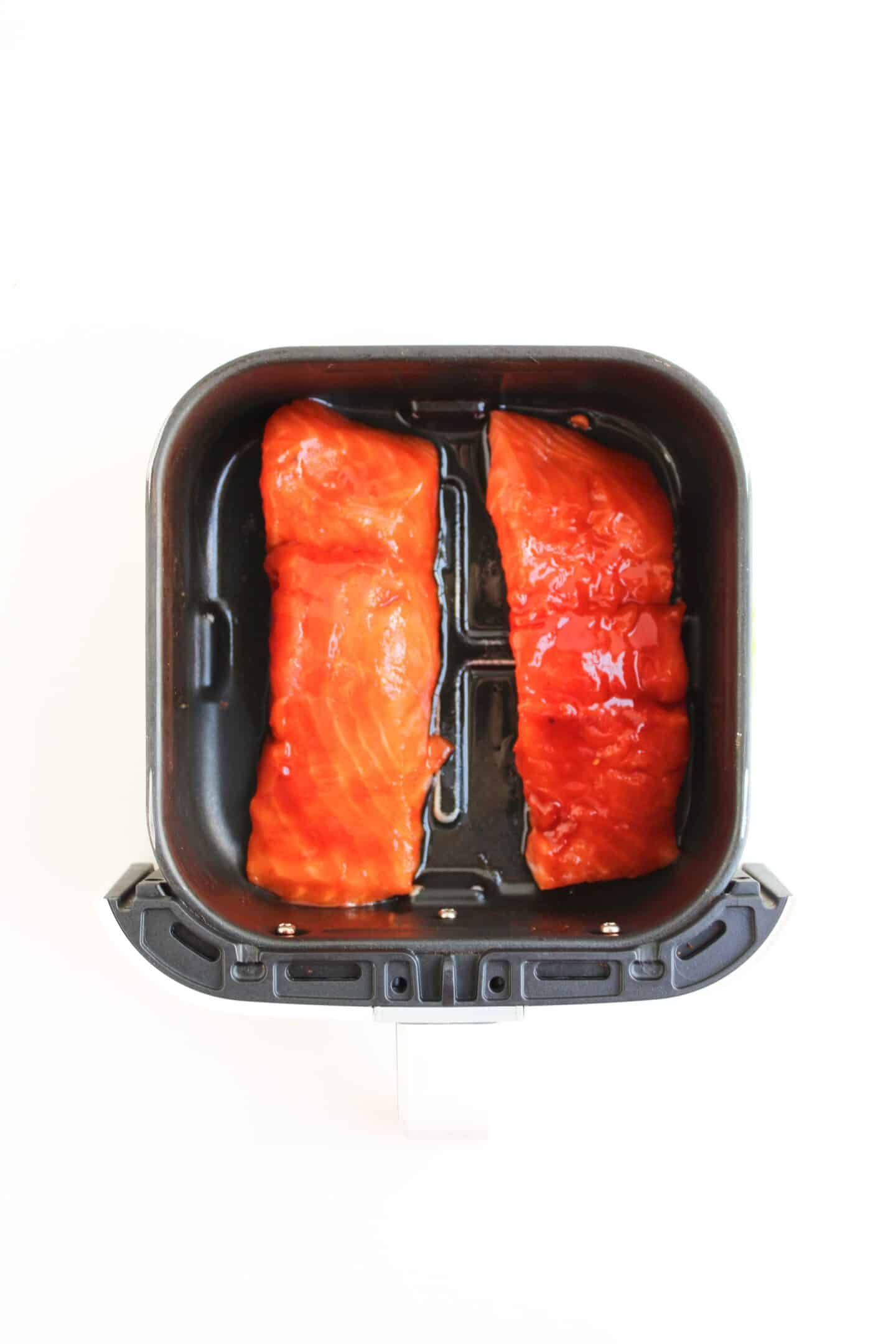 Two raw salmon fillets with a red marinade sit side by side in an air fryer basket on a white background, perfect for creating delicious Spicy Salmon Bowls or adding to your favorite Salmon Recipes.