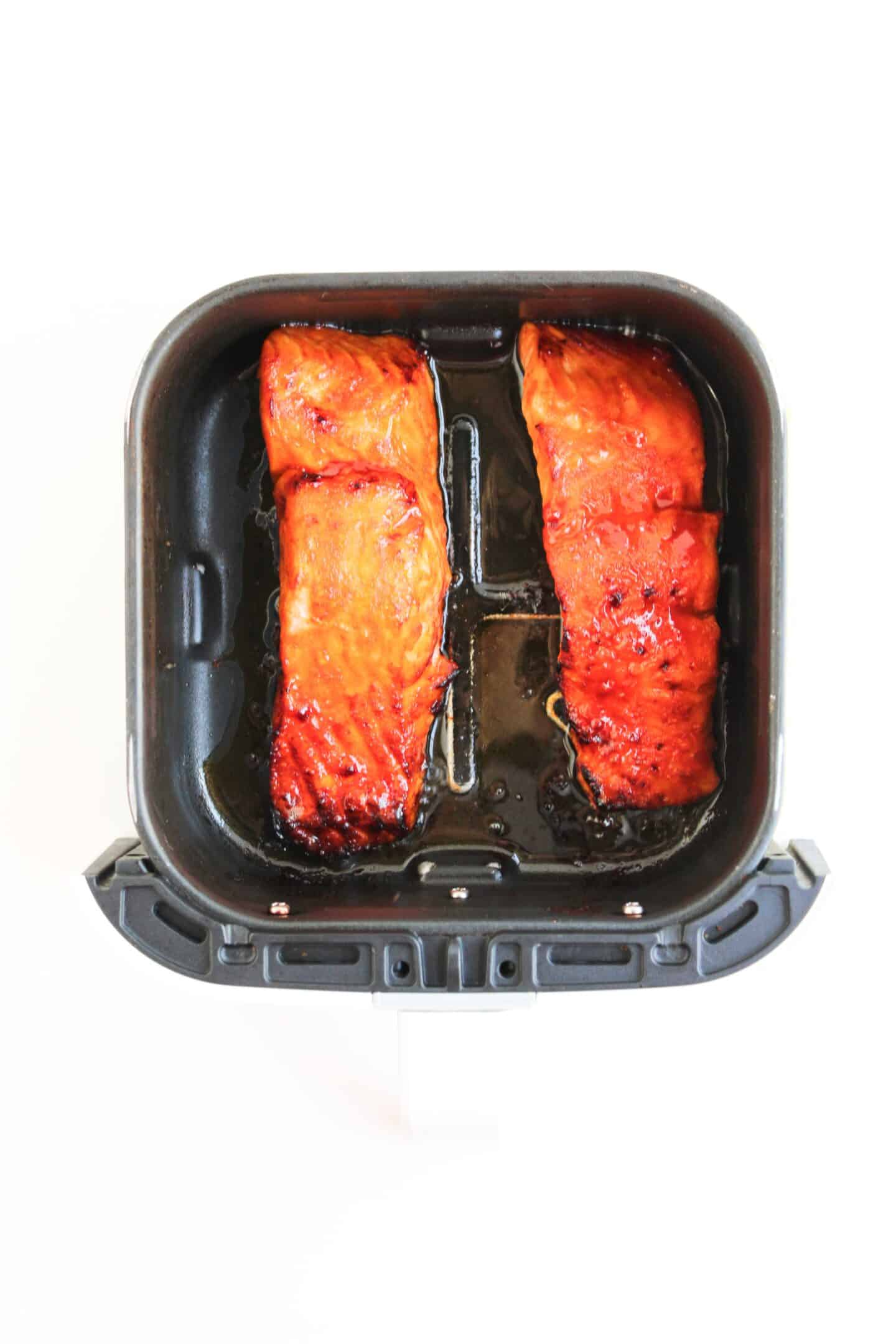 Two cooked spicy salmon fillets with a glazed surface rest inside an air fryer basket, viewed from above on a white background—perfect for adding to your favorite salmon bowls.