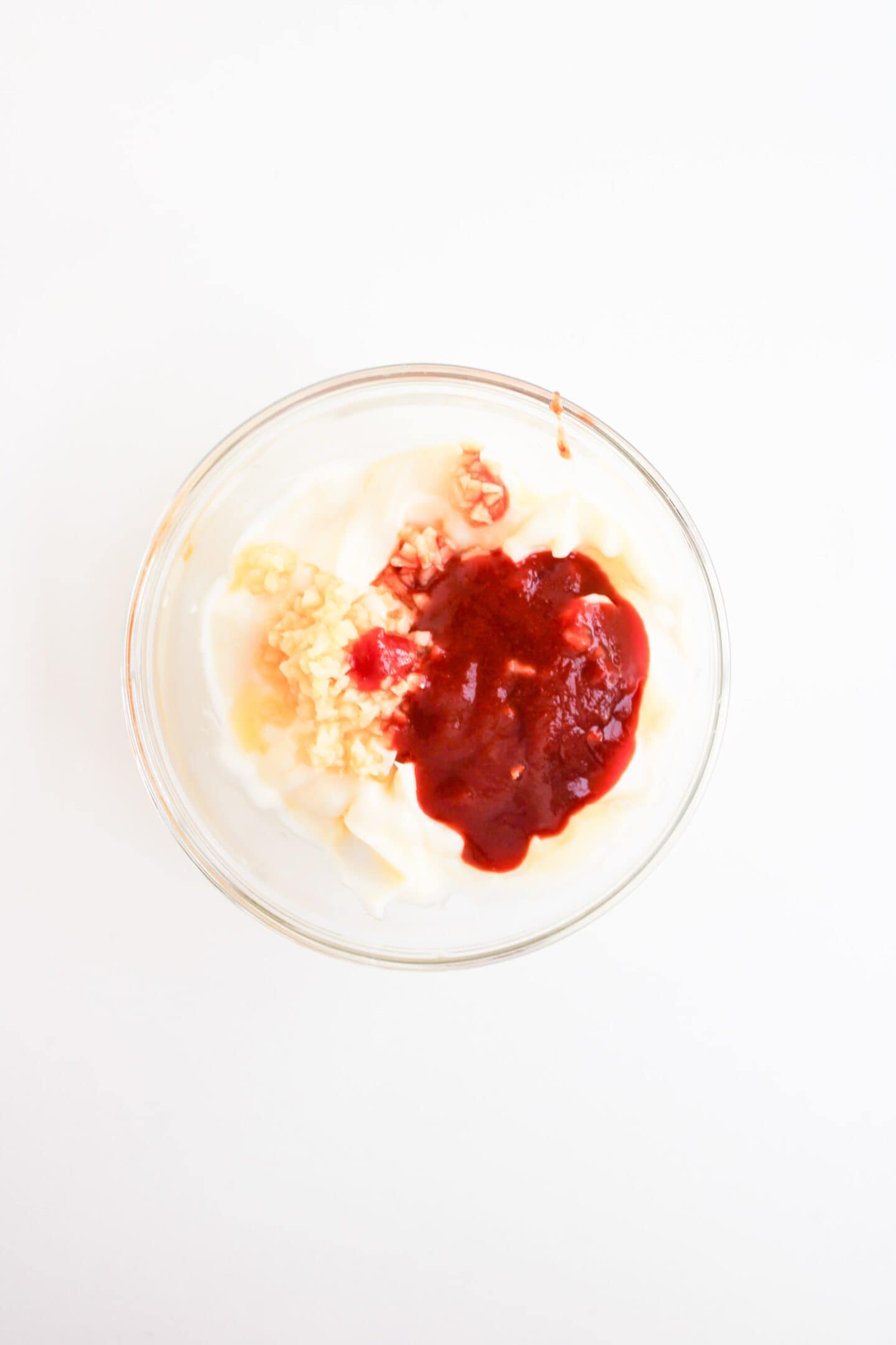 A glass bowl containing plain yogurt topped with chopped garlic and red sauce, reminiscent of the vibrant flavors found in Spicy Salmon Bowls, viewed from above on a white background.