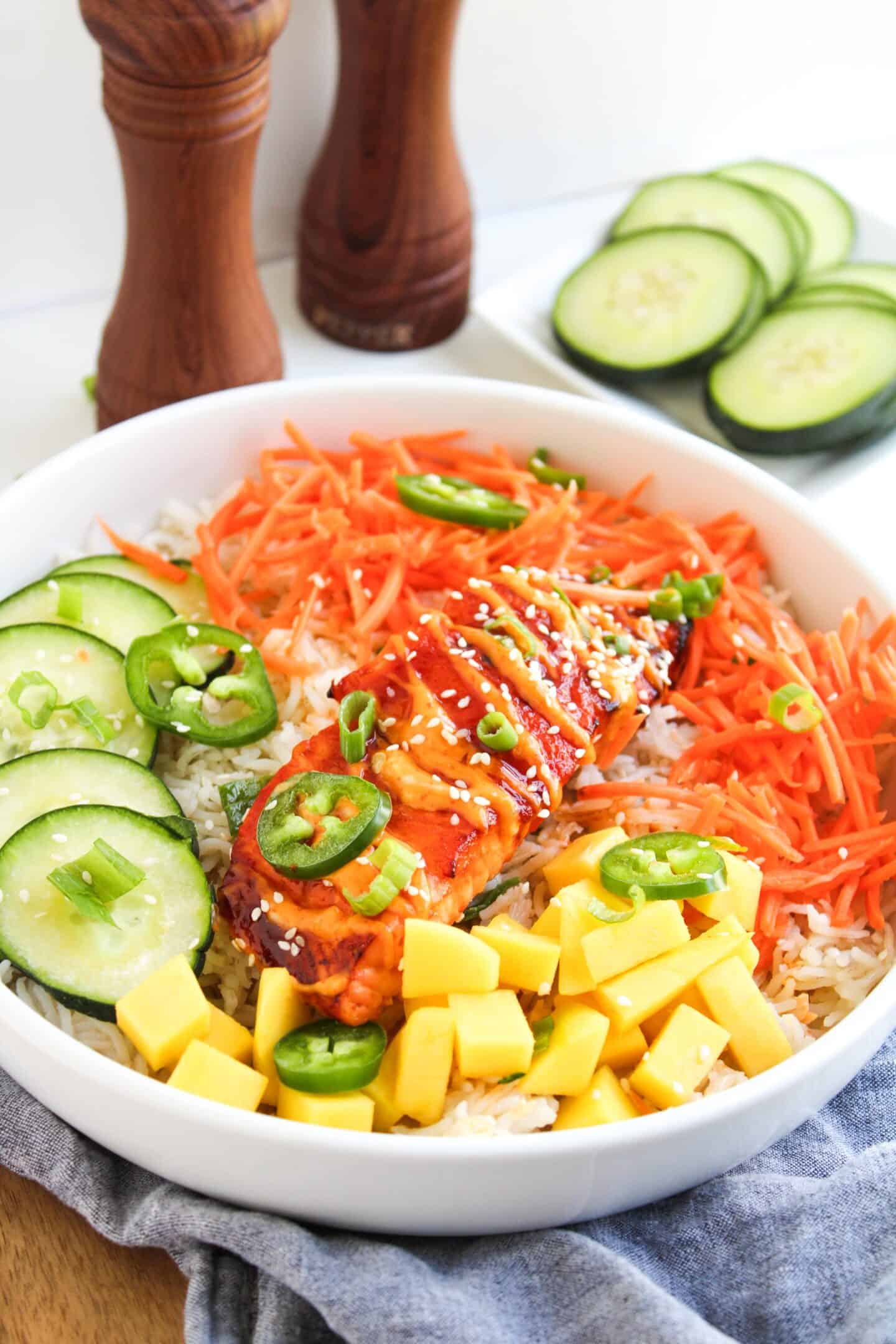 A bowl of rice topped with sliced cucumber, carrot shreds, mango chunks, jalapeño, green onions, and glazed spicy salmon, garnished with sesame seeds. Pepper mills and cucumber slices in background complete these Spicy Salmon Bowls.
