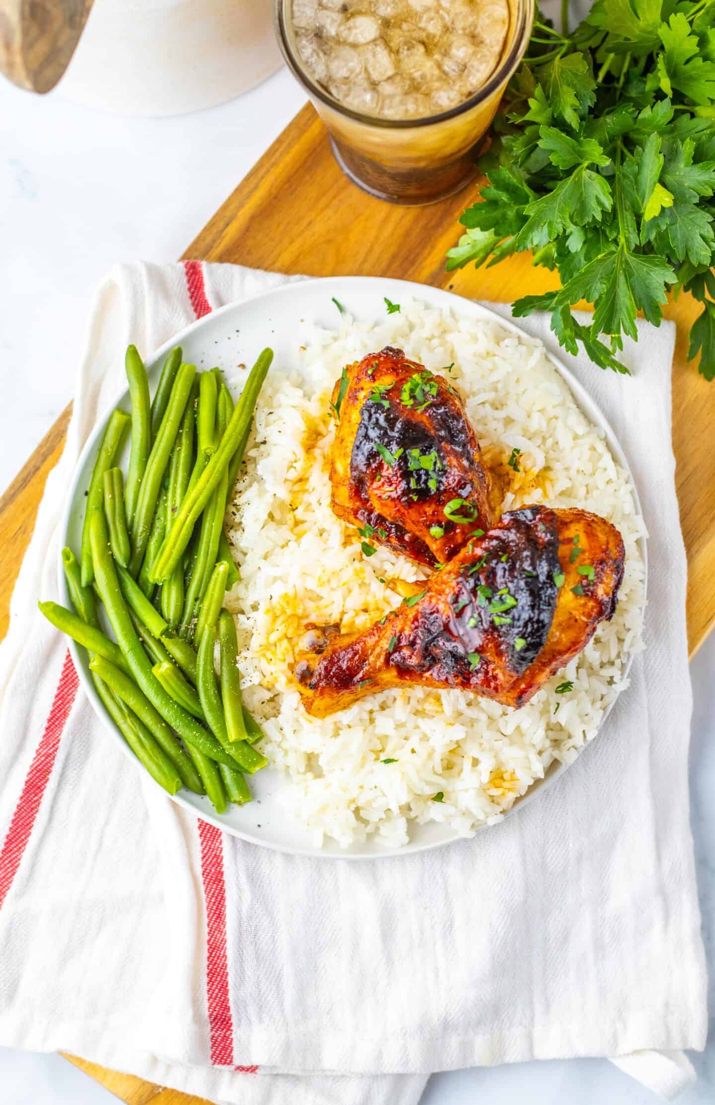 A plate with two sweet chicken legs on white rice, green beans on the side, and a drink with ice in the background, all served on a wooden board.
