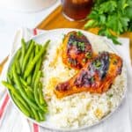 A plate with two glazed chicken legs from a sweet and smoky chicken recipe on white rice, served with green beans, next to a glass of iced beverage and fresh parsley.