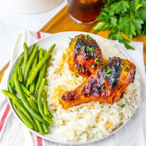 A plate with two glazed chicken legs from a sweet and smoky chicken recipe on white rice, served with green beans, next to a glass of iced beverage and fresh parsley.
