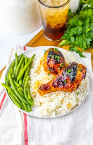 A plate with two sweet and smoky chicken legs glazed to perfection on white rice, green beans on the side, and a glass of iced beverage in the background.