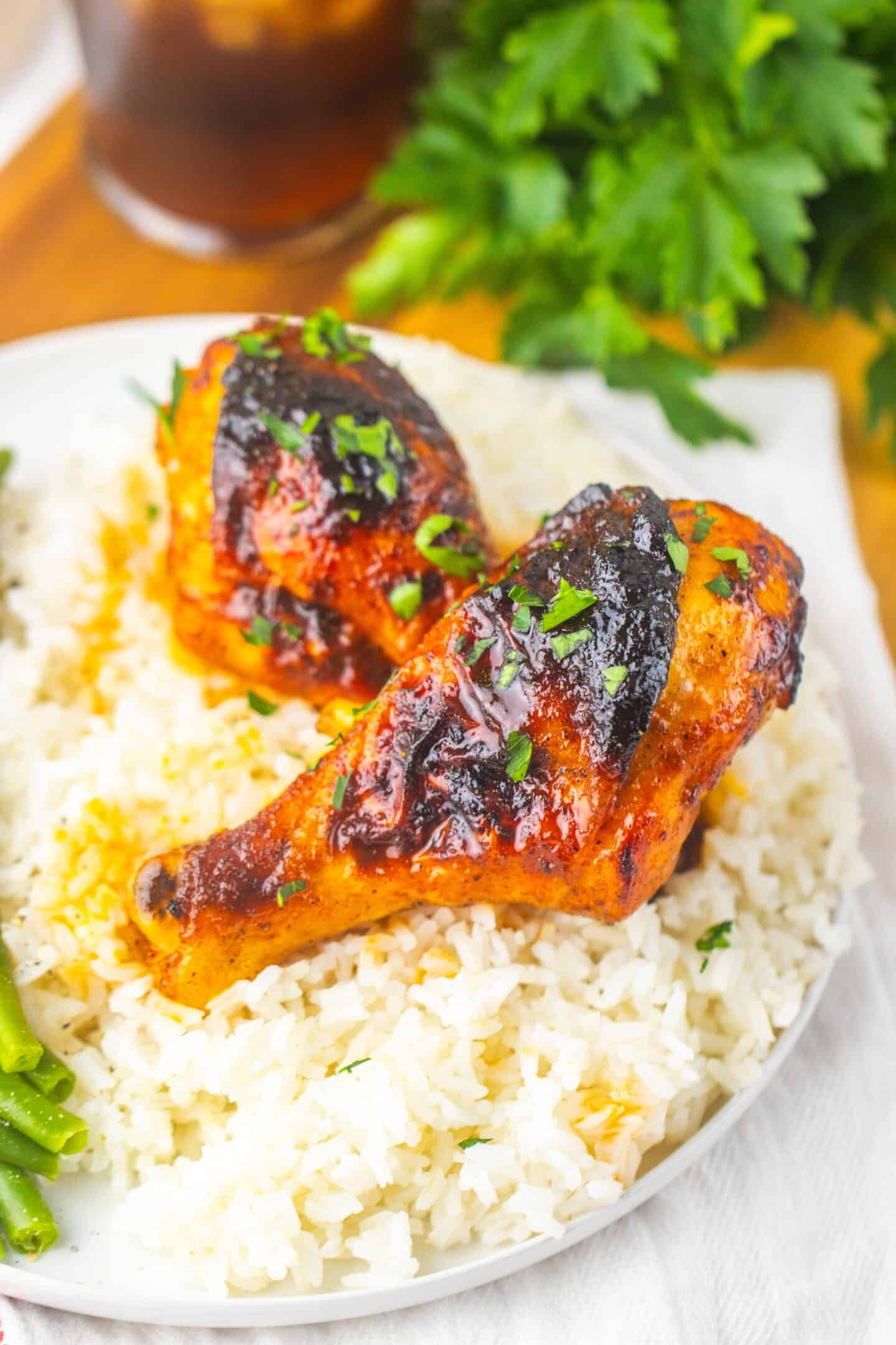 Two glazed Sweet Chicken Legs served on a bed of white rice, garnished with chopped herbs, with a side of green beans partially visible.