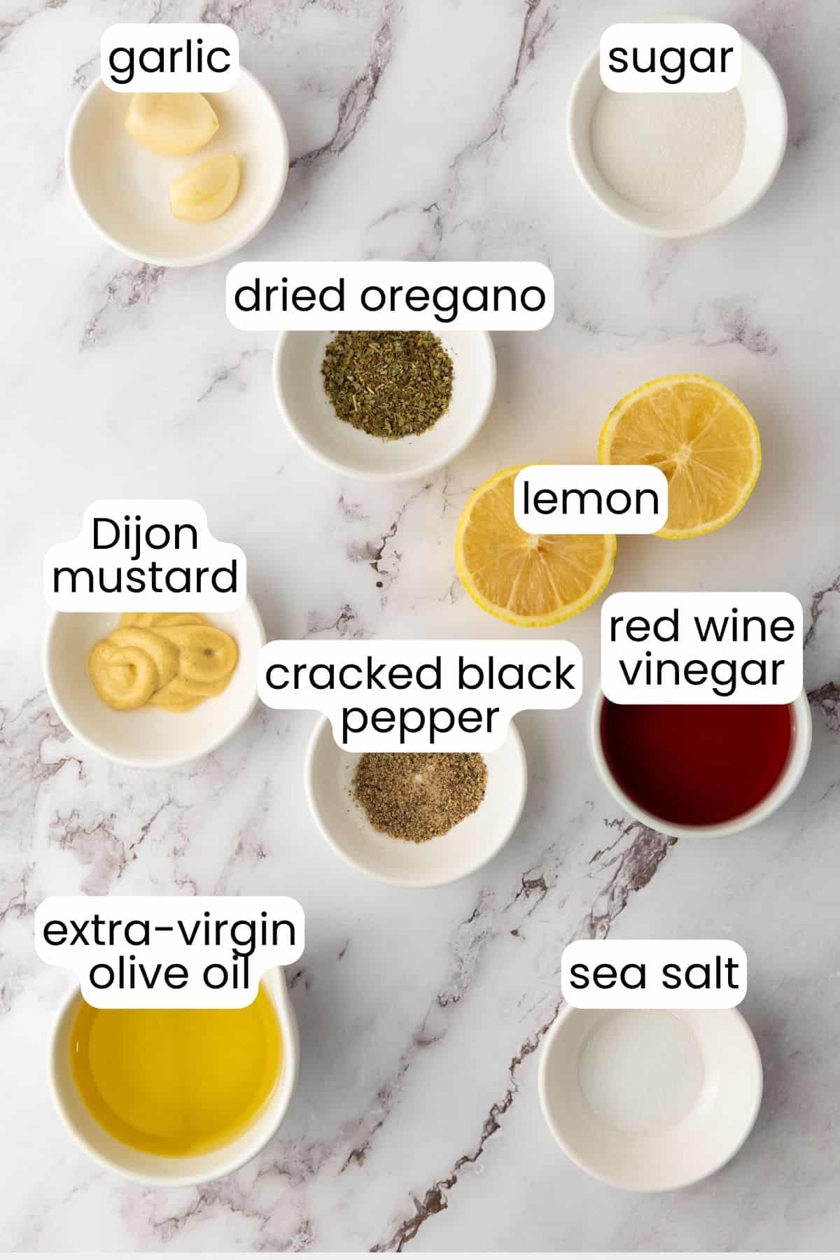 Small bowls containing garlic, dried oregano, sugar, lemon, Dijon mustard, cracked black pepper, red wine vinegar, extra-virgin olive oil, and sea salt rest on a marble surface—perfect for creating a zesty dressing for your Italian Salad.