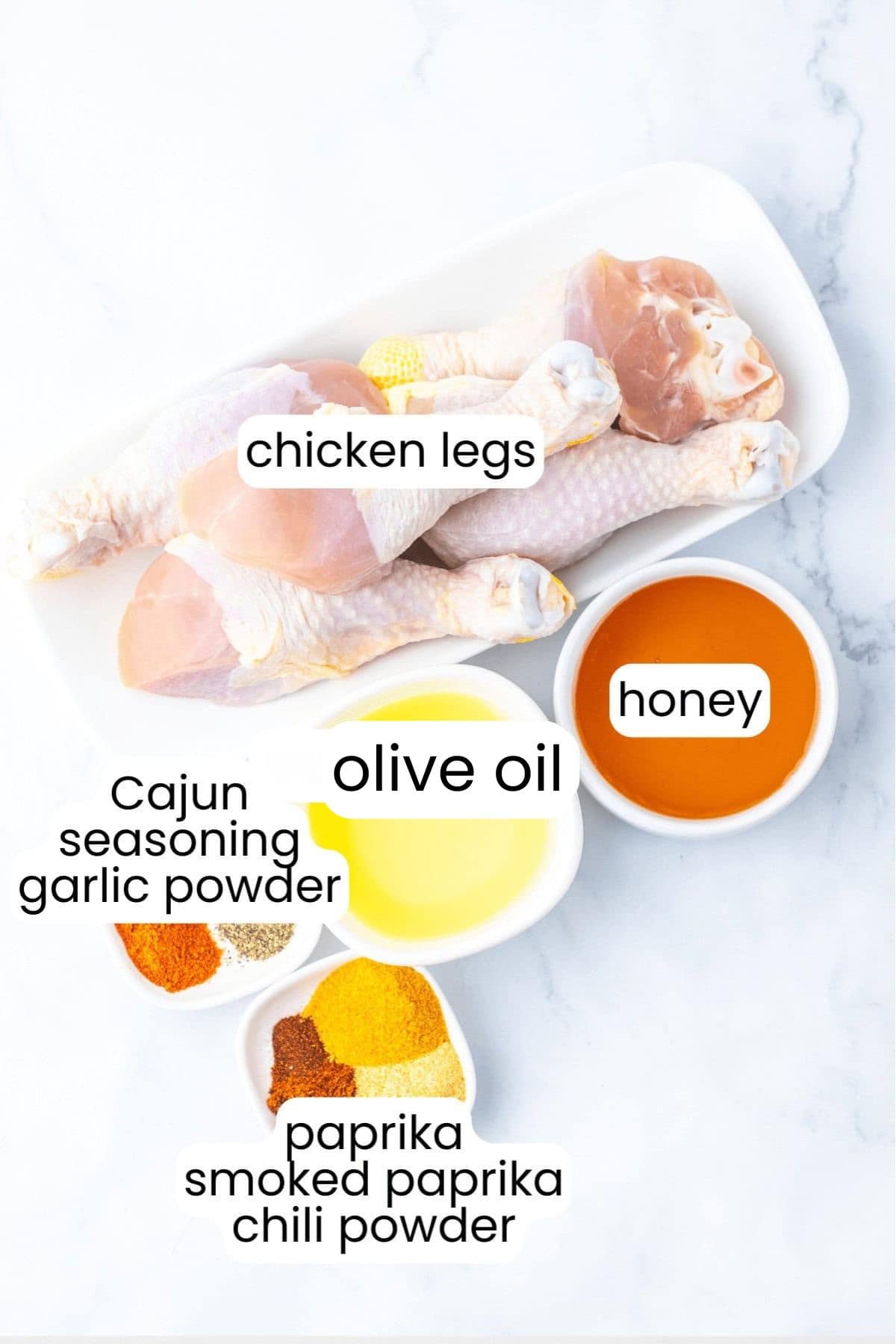 Sweet And Smoky Chicken Legs: Raw chicken legs in a dish, surrounded by bowls of olive oil and honey, plus piles of Cajun seasoning, garlic powder, paprika, smoked paprika, and chili powder on a white surface.