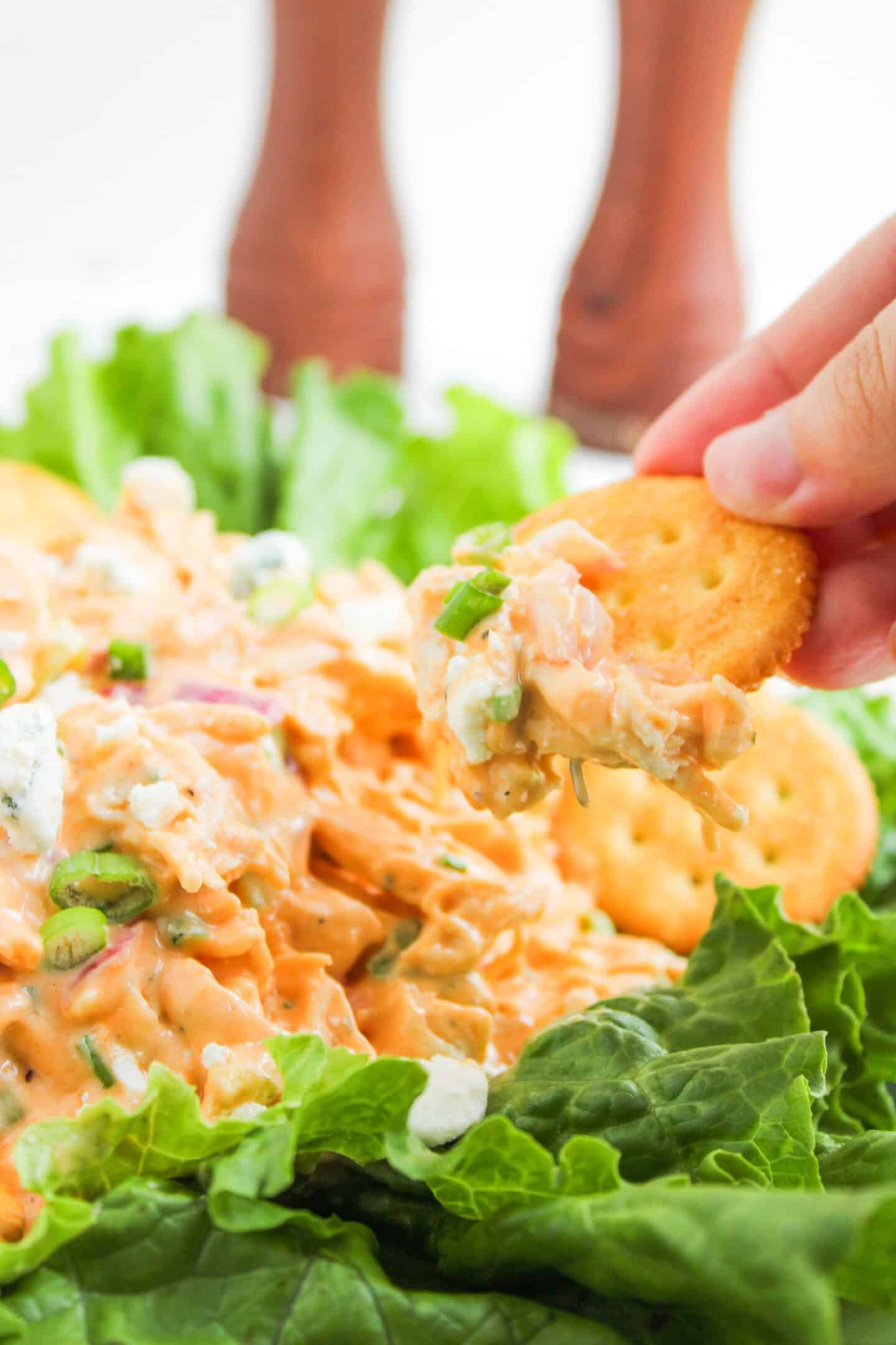 A hand holds a cracker topped with creamy Buffalo Chicken Salad dip and green onions, surrounded by lettuce leaves and more crackers.