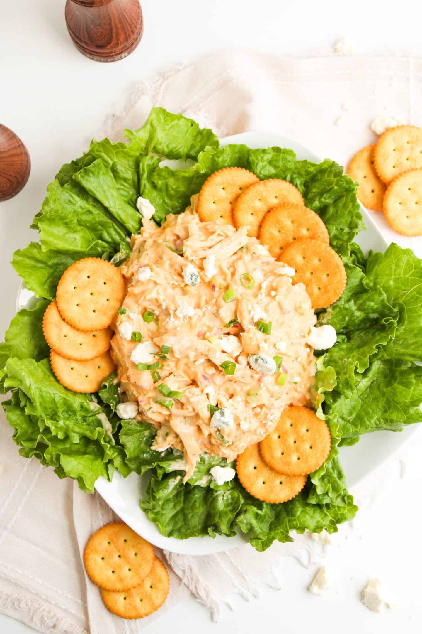 A plate of creamy Buffalo Chicken Salad topped with chopped green onions, surrounded by lettuce leaves and round crackers on a white surface.