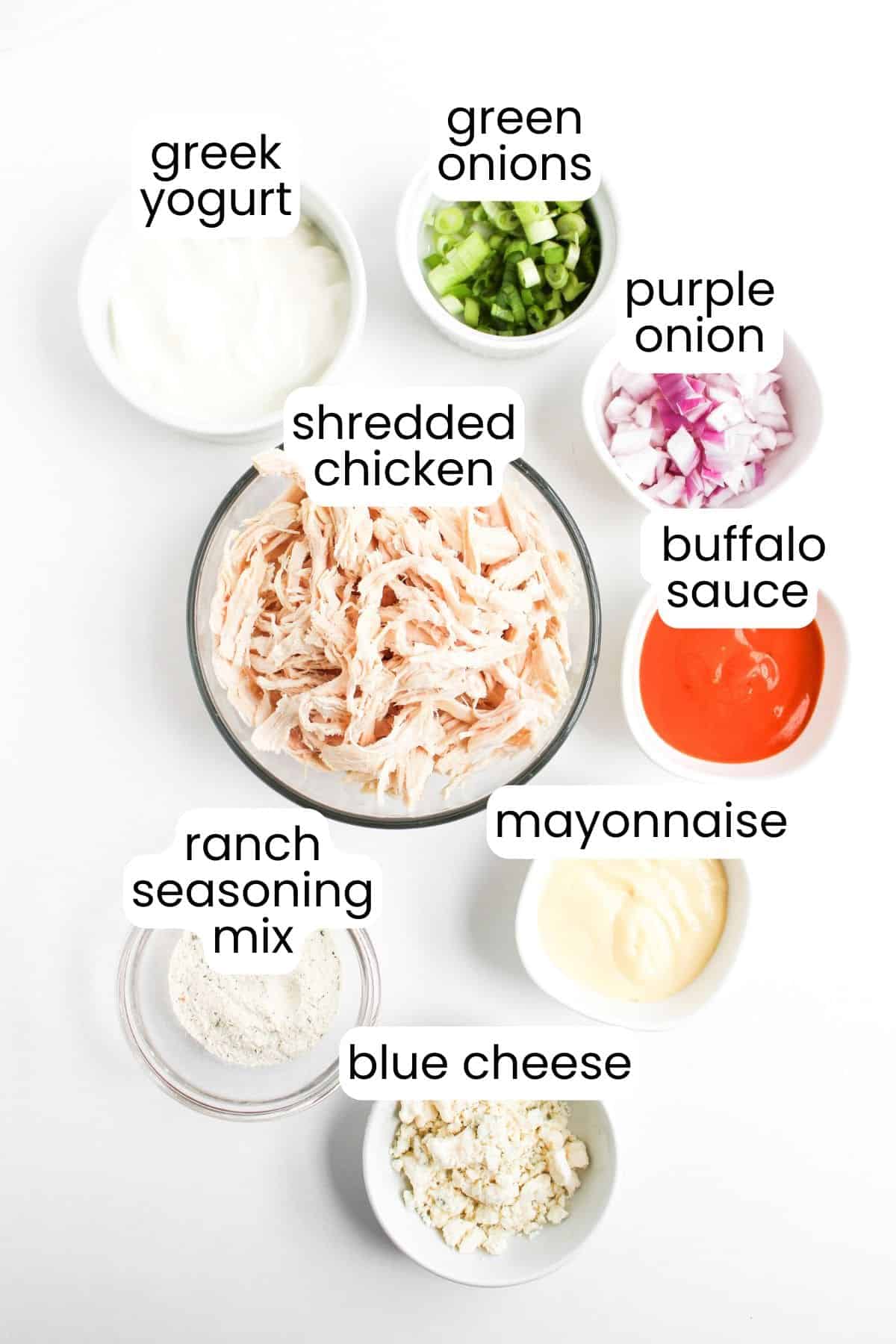 A top-down view of small bowls containing ingredients for Buffalo Chicken Salad: Greek yogurt, green onions, purple onion, shredded chicken, buffalo sauce, mayonnaise, ranch seasoning mix, and blue cheese.