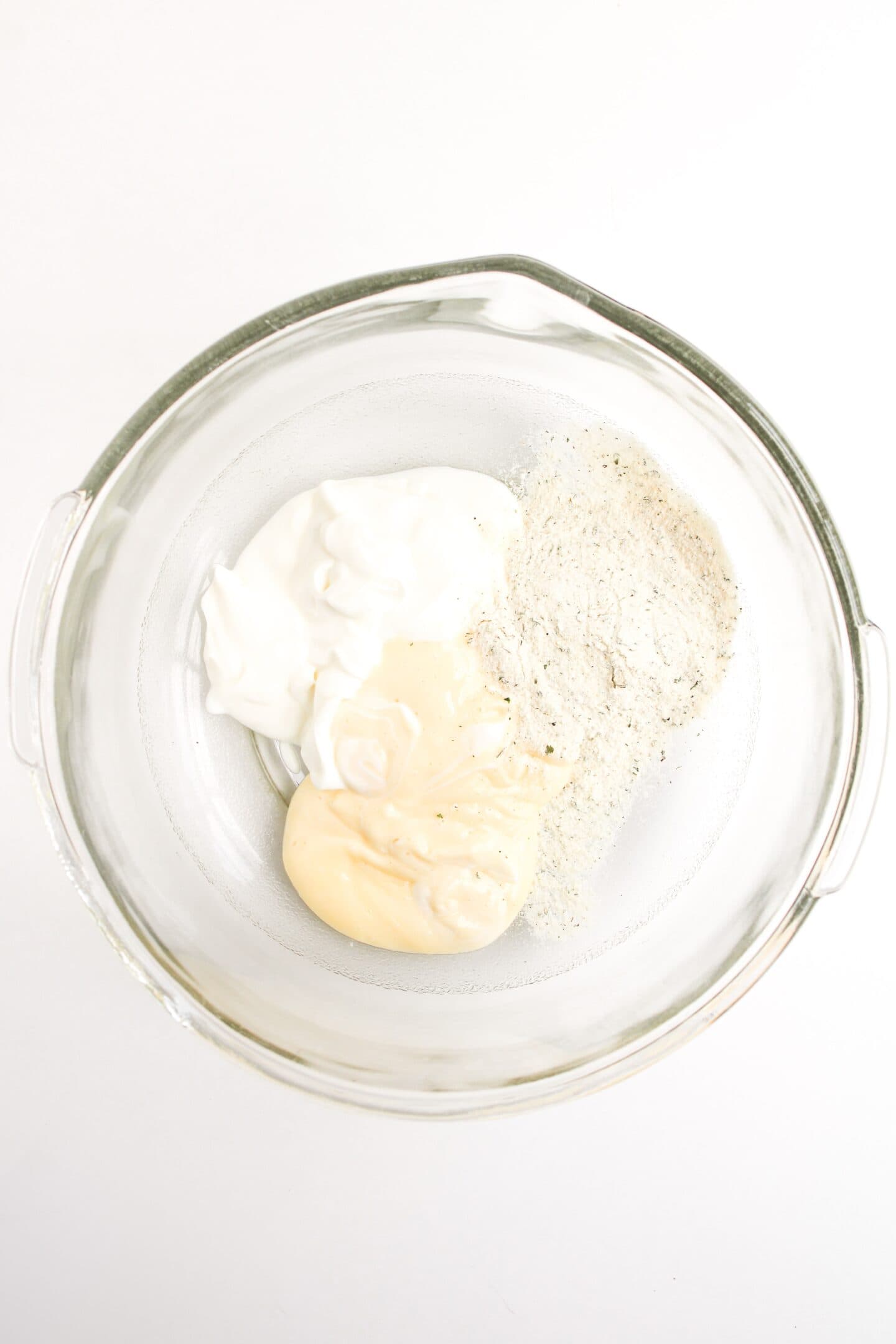 A glass bowl containing sour cream, mayonnaise, and a pile of dry seasoning mix—perfect ingredients to start your Buffalo Chicken Salad—on a white background.