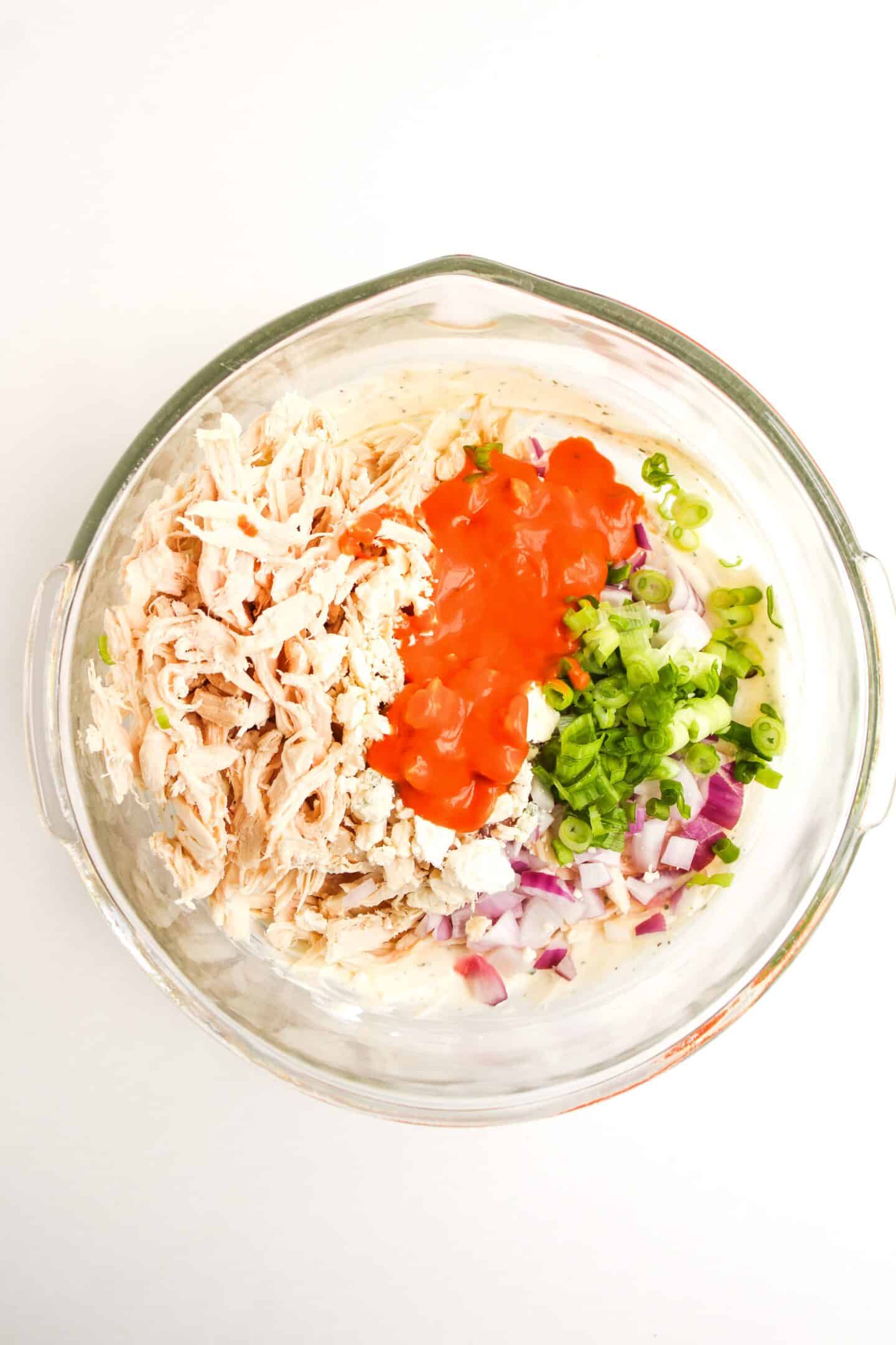 A glass bowl containing shredded chicken, buffalo sauce, chopped red onion, and sliced green onions is shown from above—creating a vibrant Buffalo Chicken Salad perfect for any meal.