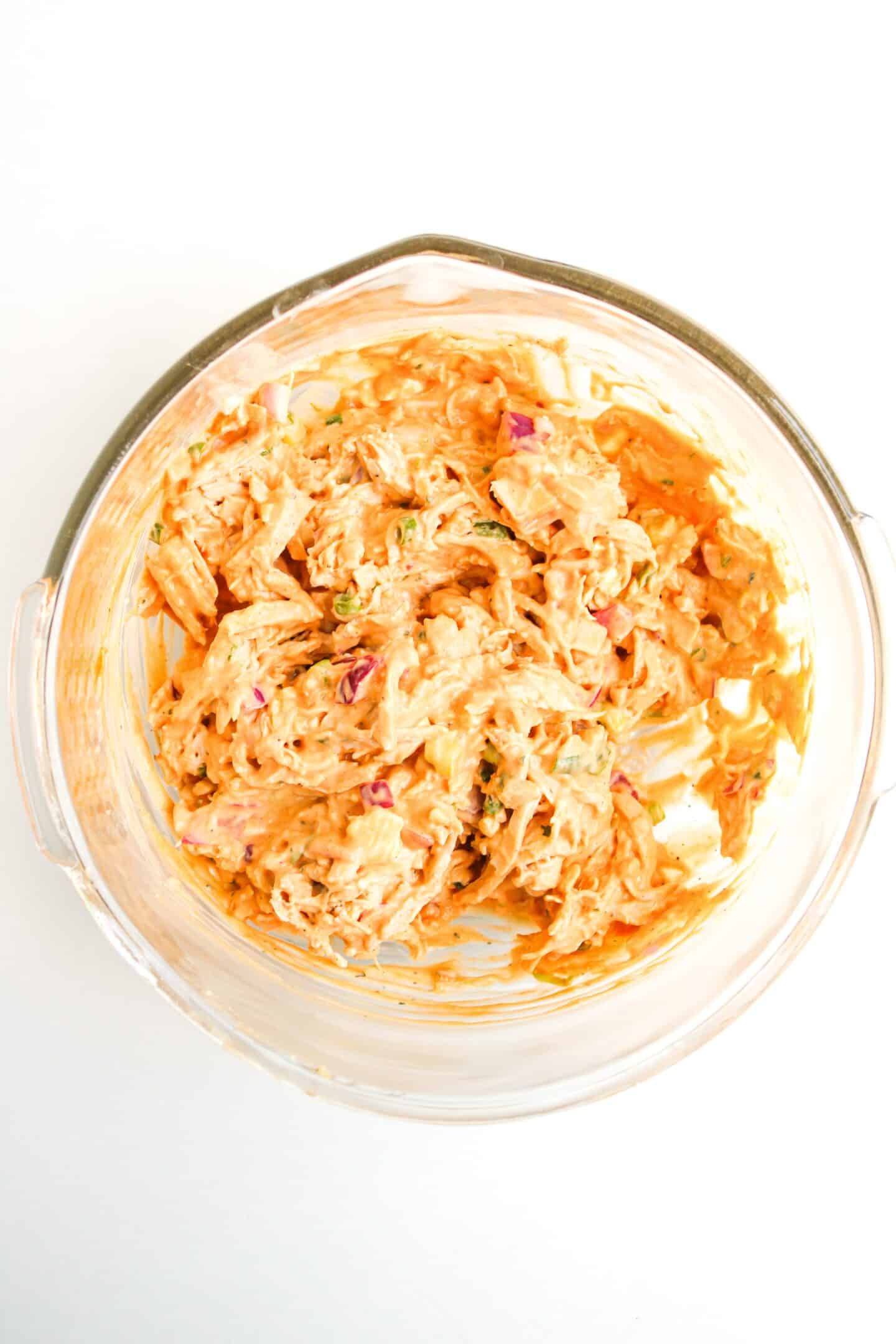 A glass bowl containing a creamy Buffalo Chicken Salad with visible herbs and red onions on a white background.