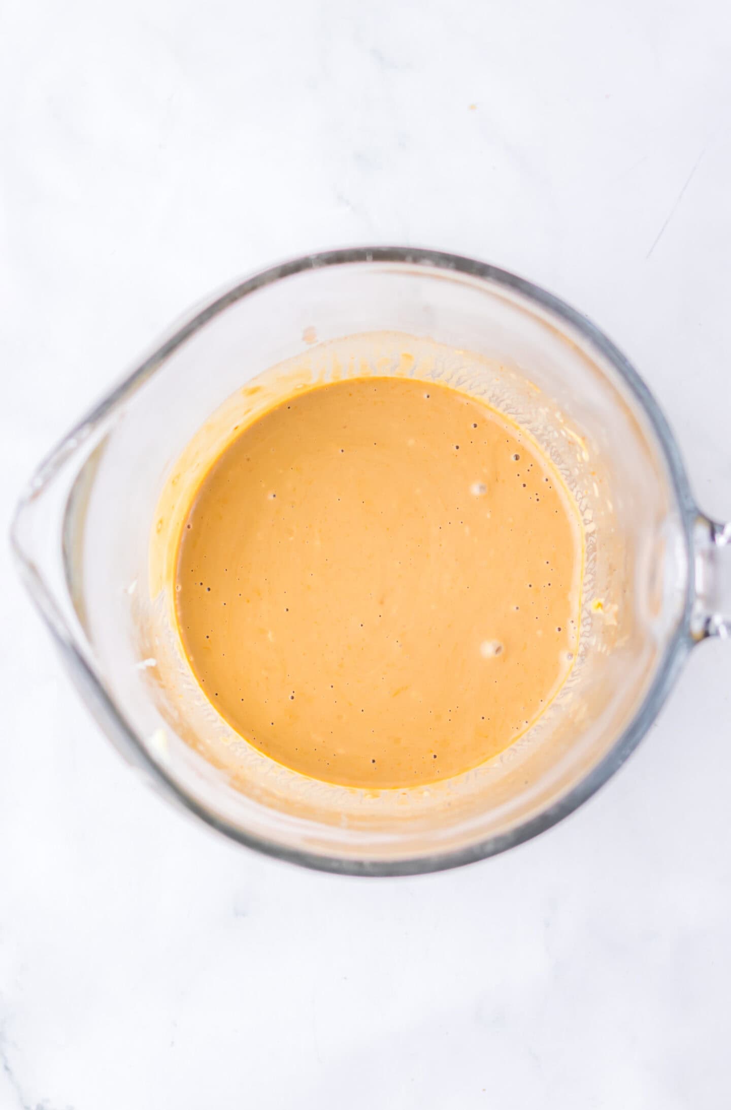A glass measuring cup filled with a smooth, light brown batter or sauce—perfect for drizzling over your Firecracker Meatballs—sits on a white surface.