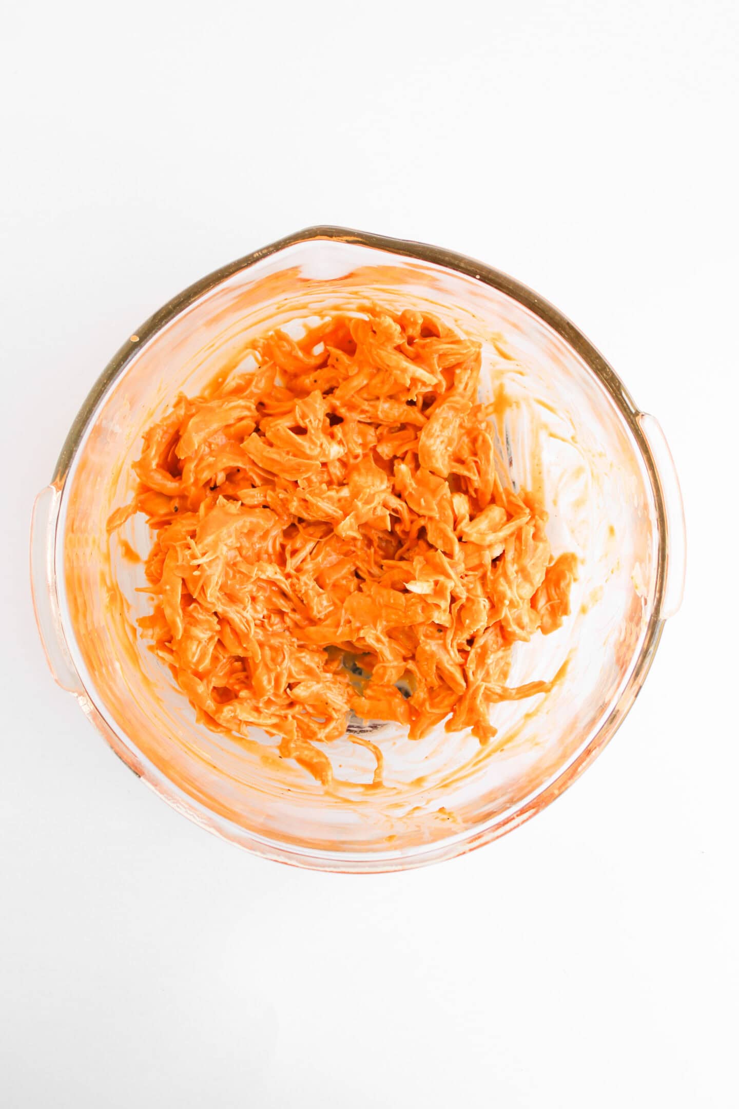 Clear glass bowl containing shredded chicken mixed with an orange sauce, perfect for topping Buffalo Chicken Stuffed Baked Potatoes, placed on a white background.