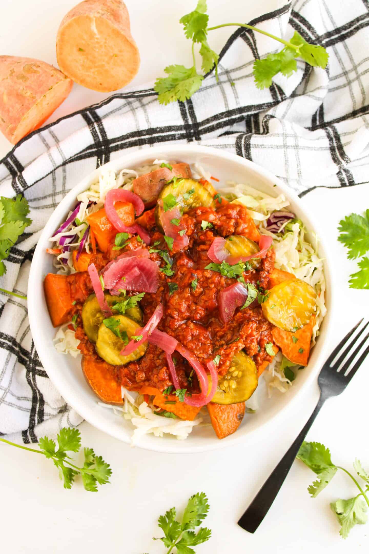 A hearty Sloppy Joe Bowl: chili topped with pickles and onions, served over shredded cabbage and sweet potatoes, garnished with cilantro and paired with a black fork on the side.