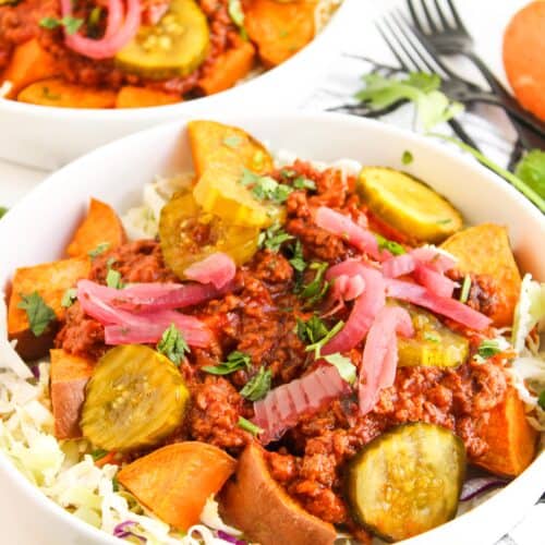 A bowl of shredded cabbage topped with chili, sliced pickles, sweet potato chunks, and pickled onions—an inventive twist on classic Sloppy Joe Bowls—with another similar bowl and utensils in the background.