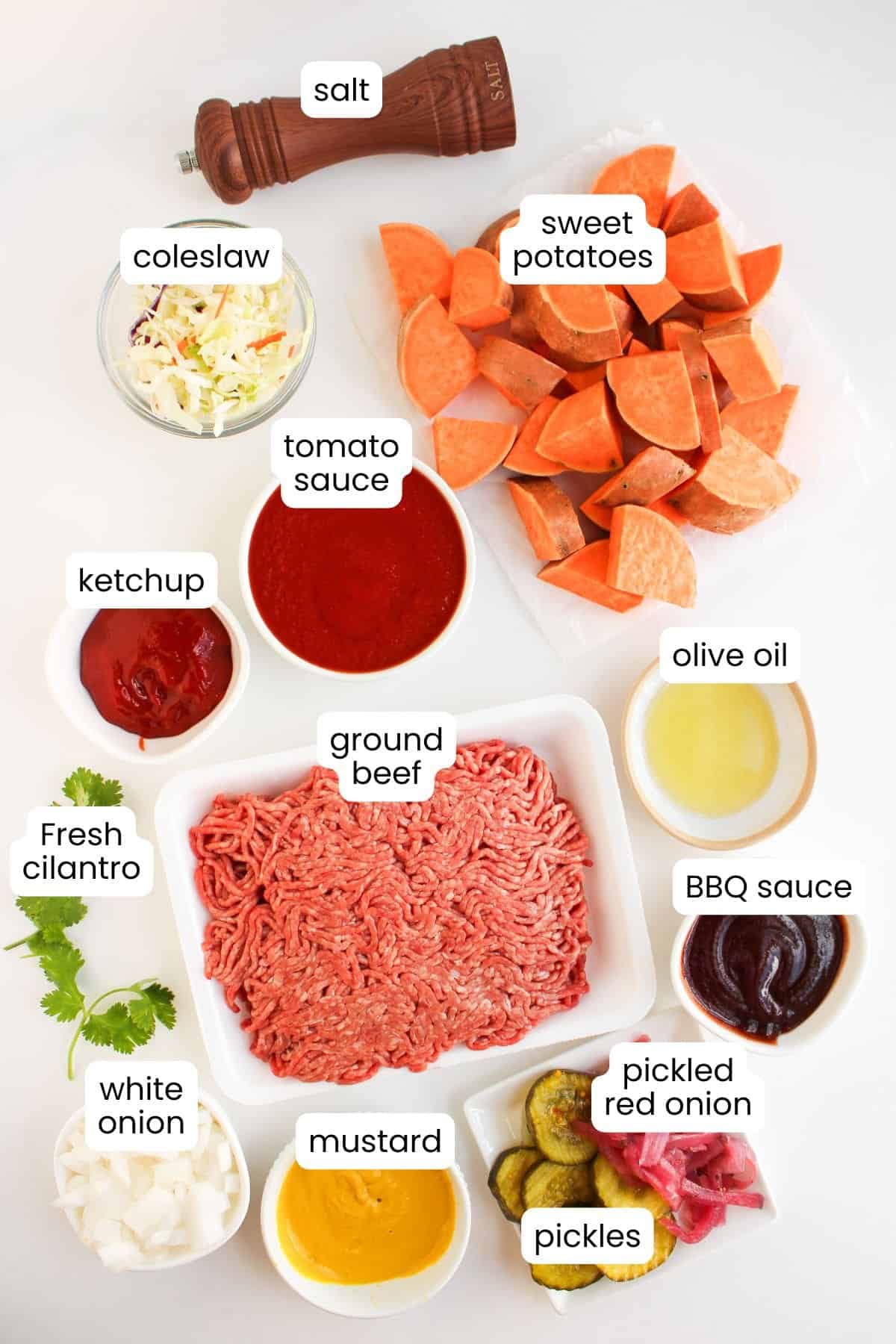 Assorted ingredients on a white surface, perfect for preparing easy dinner Sloppy Joe Bowls—includes ground beef, sweet potatoes, coleslaw, sauces, white onion, pickles, pickled red onion, fresh cilantro, olive oil, and a salt grinder.